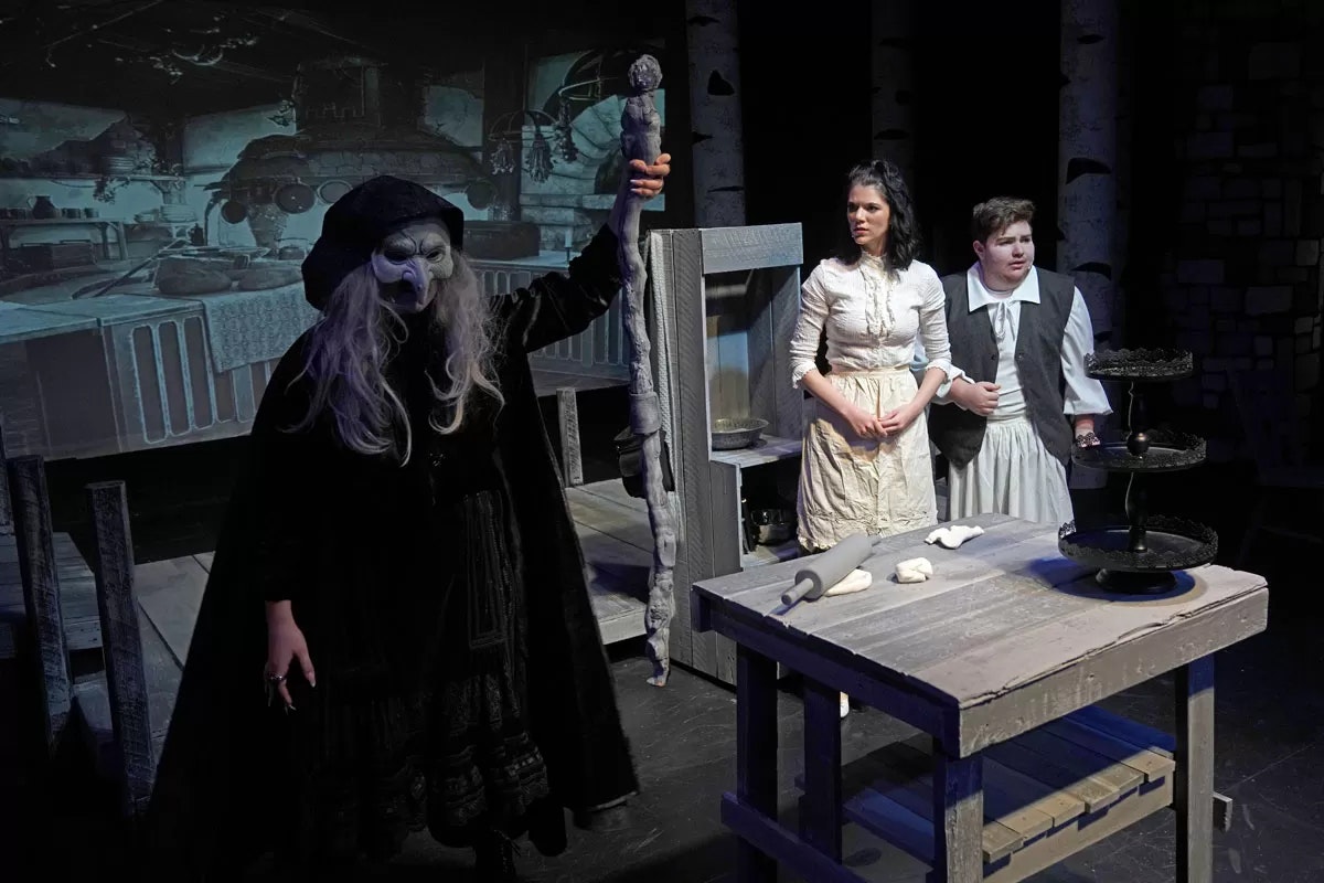 A theatrical scene with a masked figure holding a staff, two other characters, and a table with dough and rolling pin.