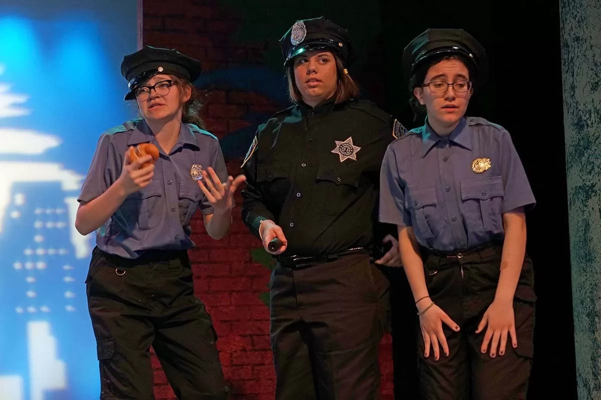 Three people in police uniforms, one holding a donut, on stage with a brick wall and blue lighting in the background.
