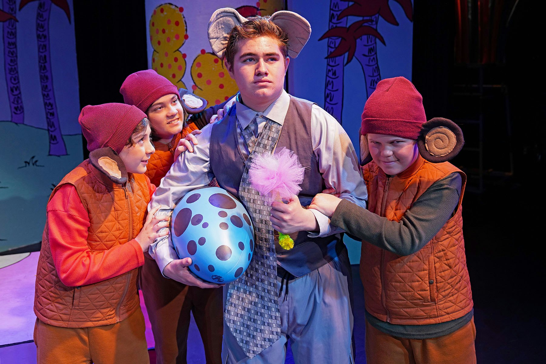A group of children dressed in costumes interacts in a playful scene, featuring an elephant-themed character holding a blue egg.
