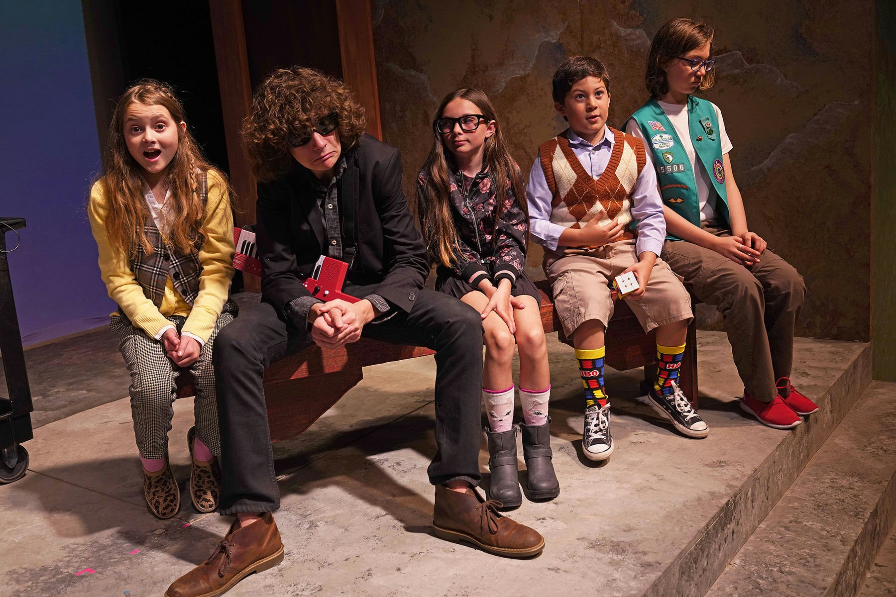 A group of kids in quirky outfits sits on a bench, showcasing a mix of fashion styles and expressions in a playful setting.