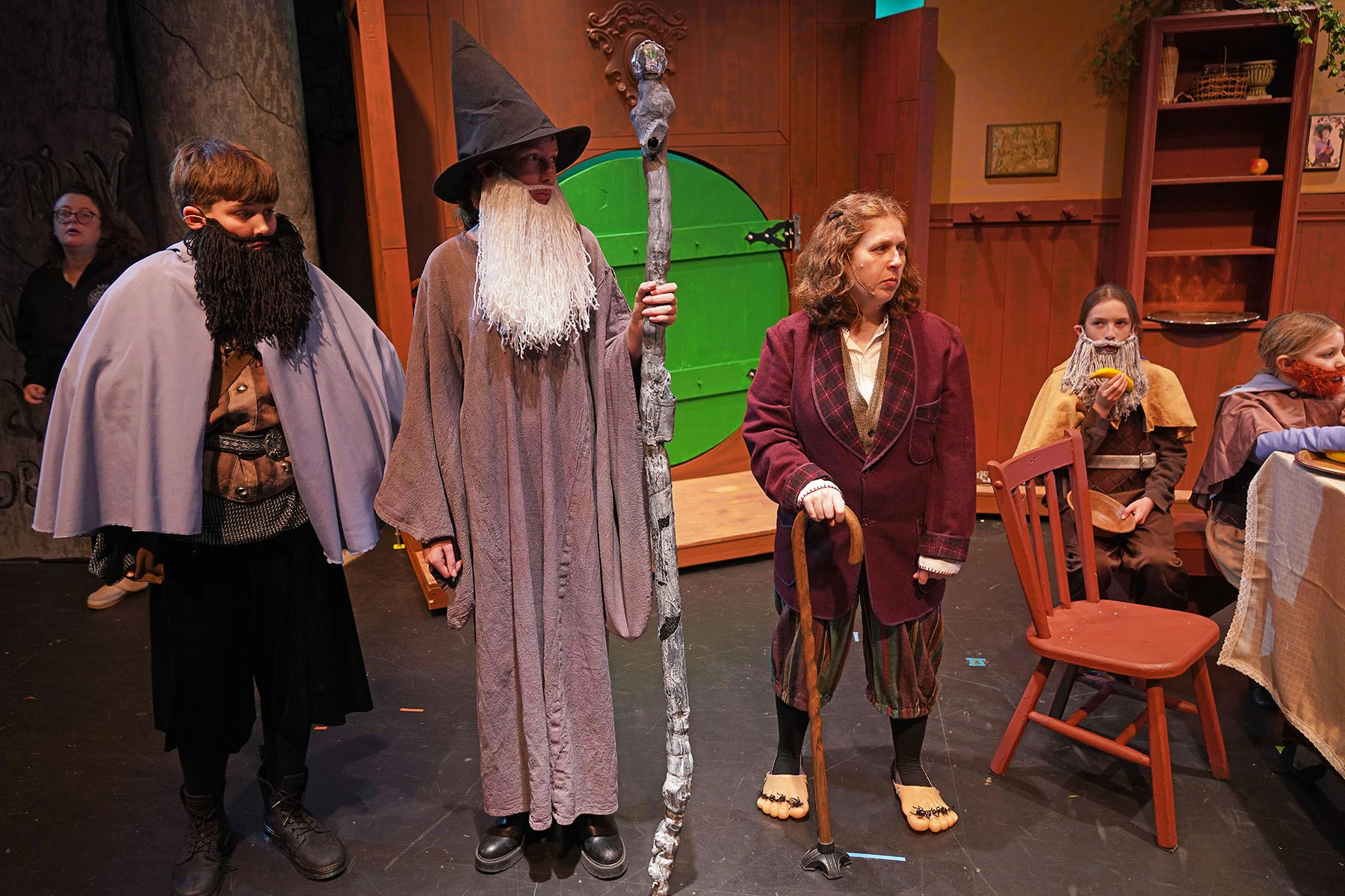 A group of people in fantasy costumes on stage, featuring a wizard, bearded characters, and a hobbit-like figure with a walking stick.