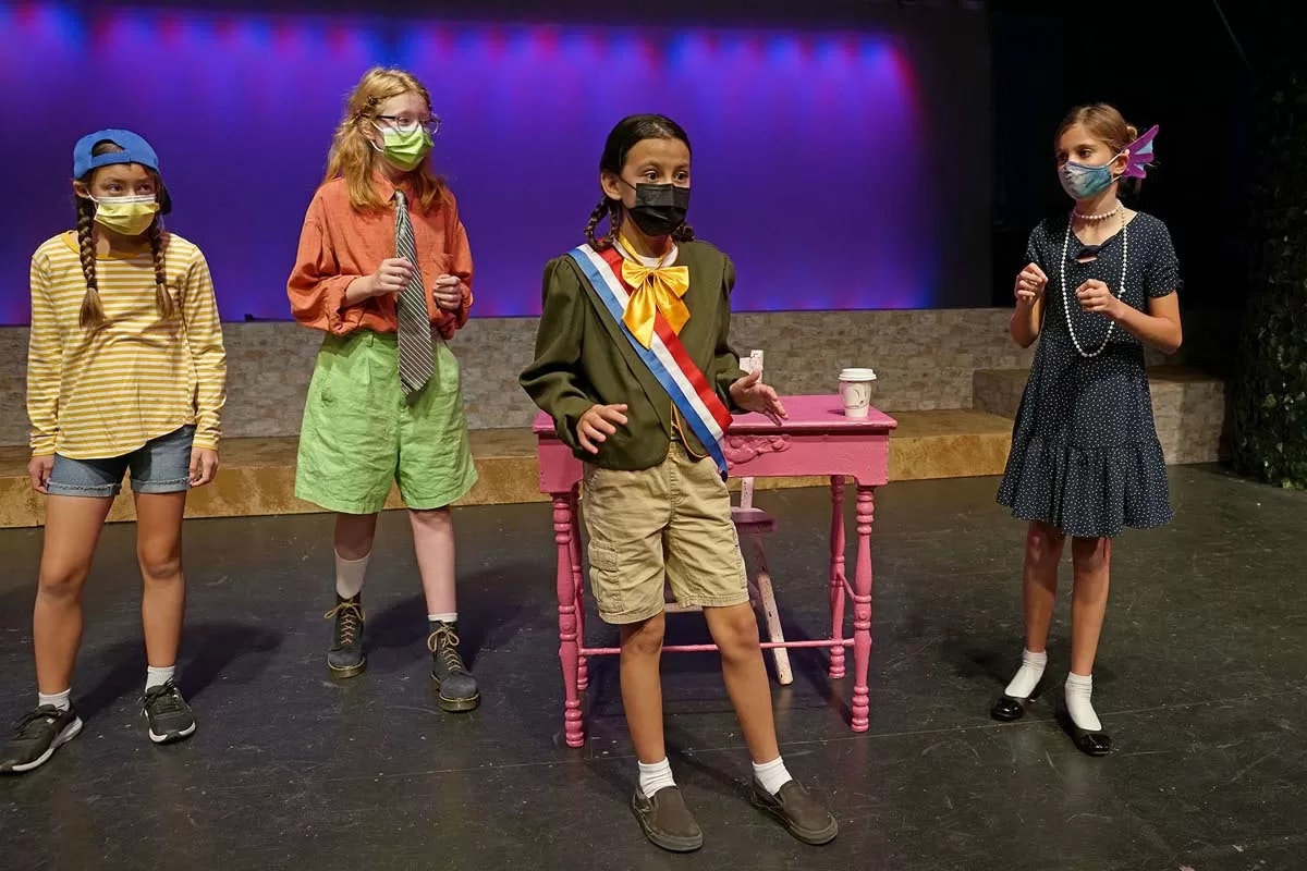 Four children in costumes and masks perform on stage with colorful lighting and a pink table.