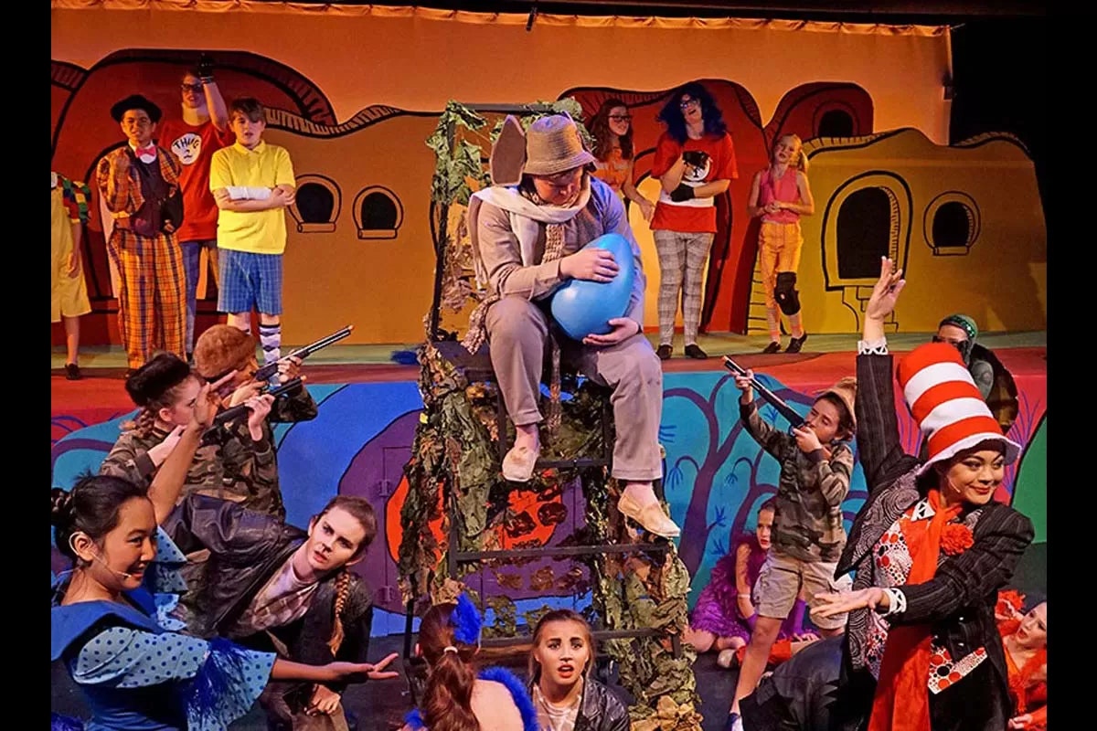 A colorful stage play scene with costumed characters, including one holding a large blue egg.