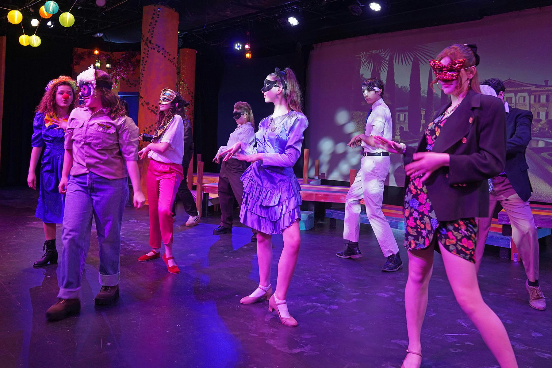 A group of masked performers on stage in colorful costumes, engaged in a dramatic scene with vibrant lighting.