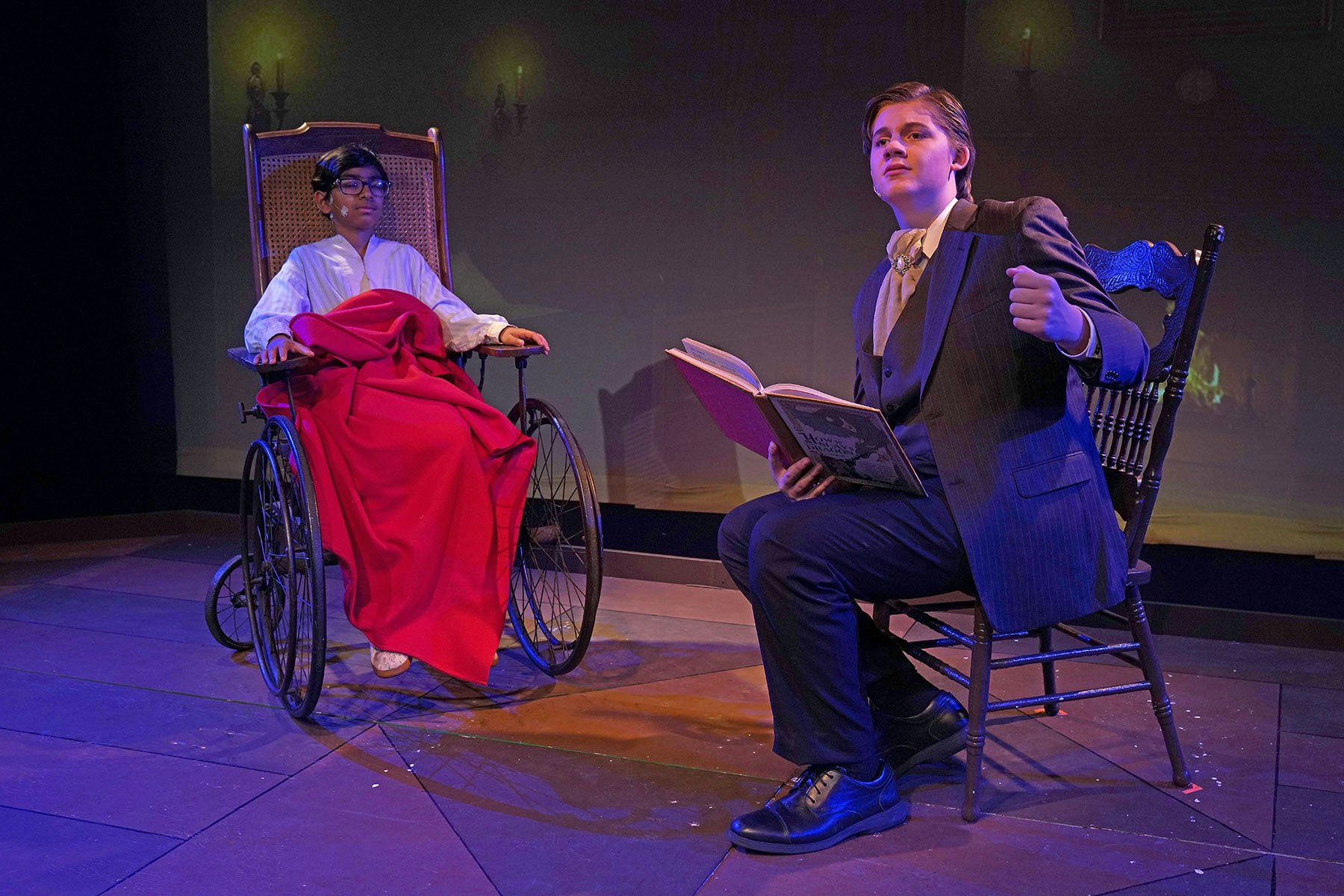 Two people on stage: one in a wheelchair with a red blanket; the other in a suit, sitting and reading a book.