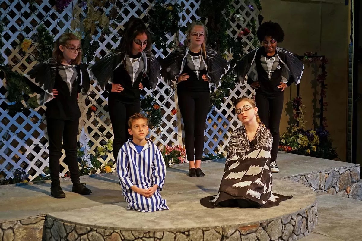 A stage scene with actors in costume; two seated in front, four standing behind dressed in dark outfits with wing-like features.