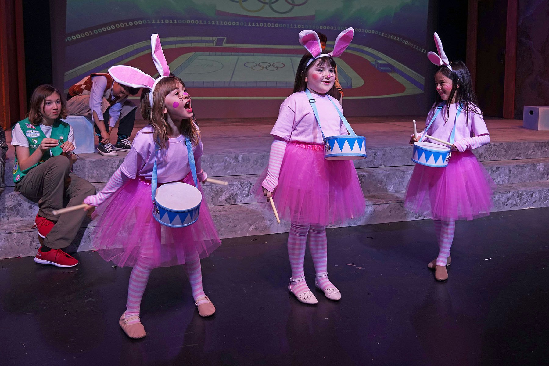 Children dressed as bunnies in pink tutus perform on stage, holding drums. One girl is singing passionately.
