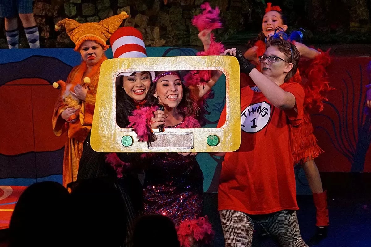 A colorful stage performance with people in costumes, including "Thing 1" and a person wearing a red and white striped hat.