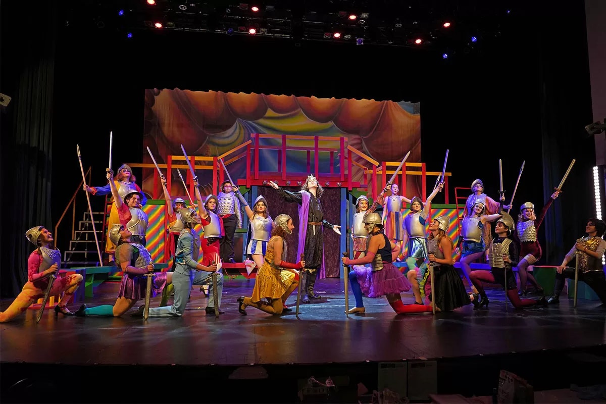 A colorful theatrical performance with actors in vibrant costumes holding staffs, set on a bright stage with dramatic lighting.