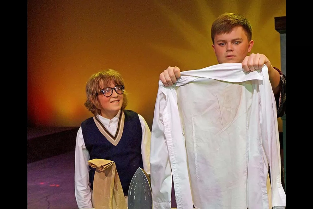 Two people, one holding a wrinkled shirt, the other smiling, with an iron and paper bag nearby, in a theater setting.