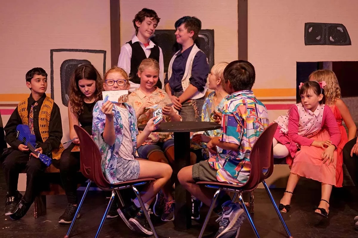A group of children in colorful outfits are sitting and interacting on stage, possibly during a theatrical performance.