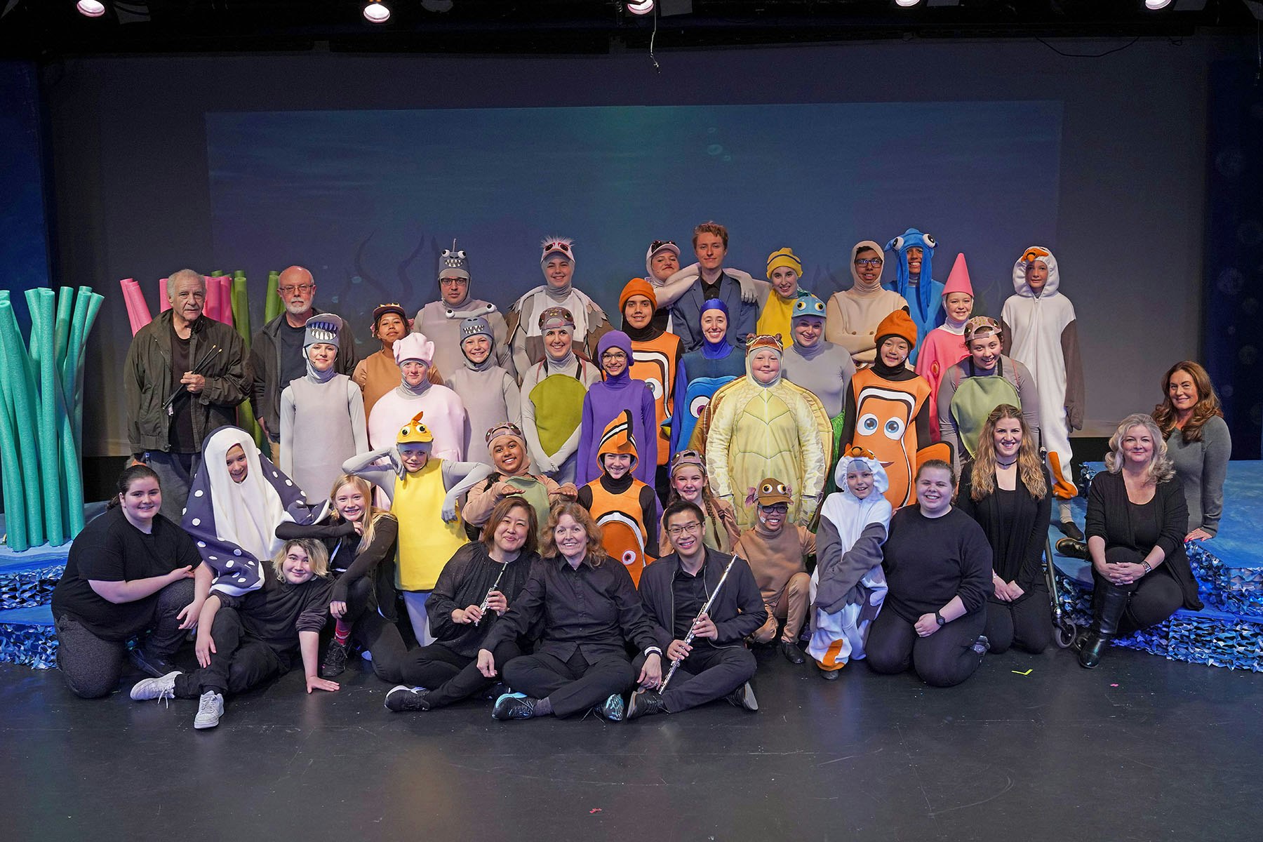 A group dressed in colorful aquatic-themed costumes poses together on a stage set resembling an underwater scene.