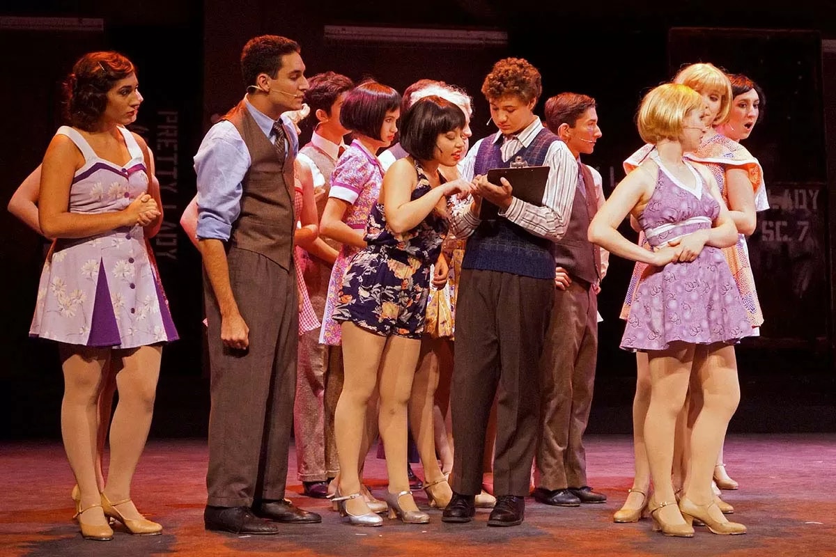 A group of performers in colorful costumes stand together on stage, with one person holding a clipboard.