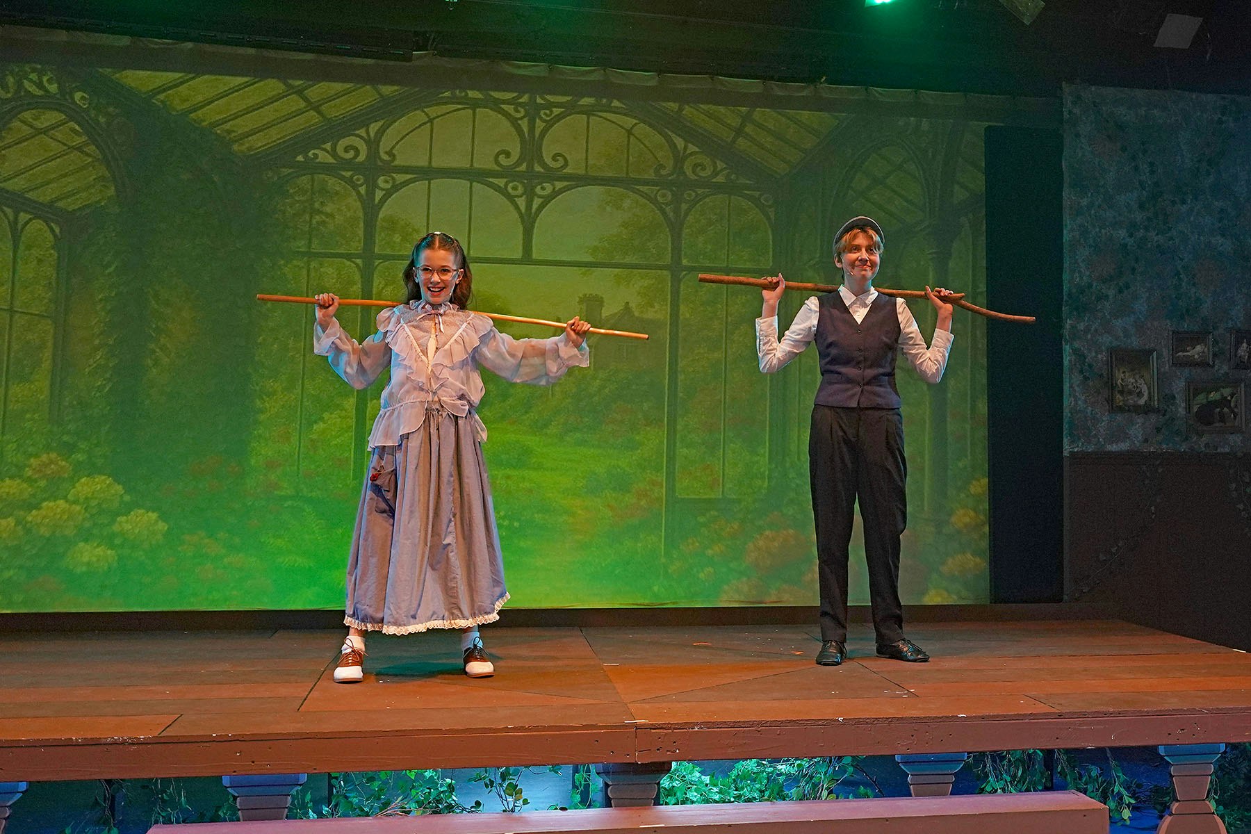 Two people on stage holding wooden rods, with a scenic backdrop of a garden conservatory.