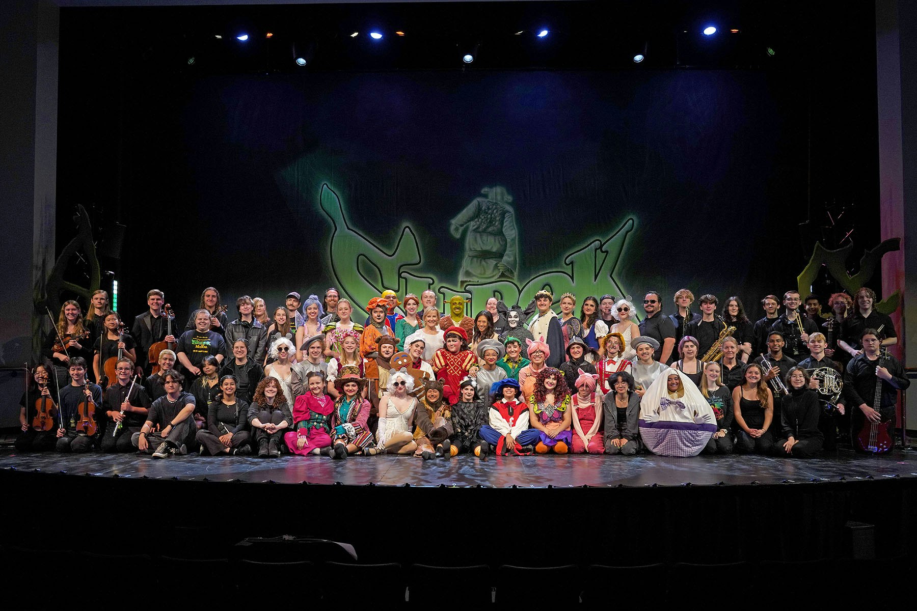 A large group of people in colorful costumes on stage with "Shrek" backdrop, some holding musical instruments.