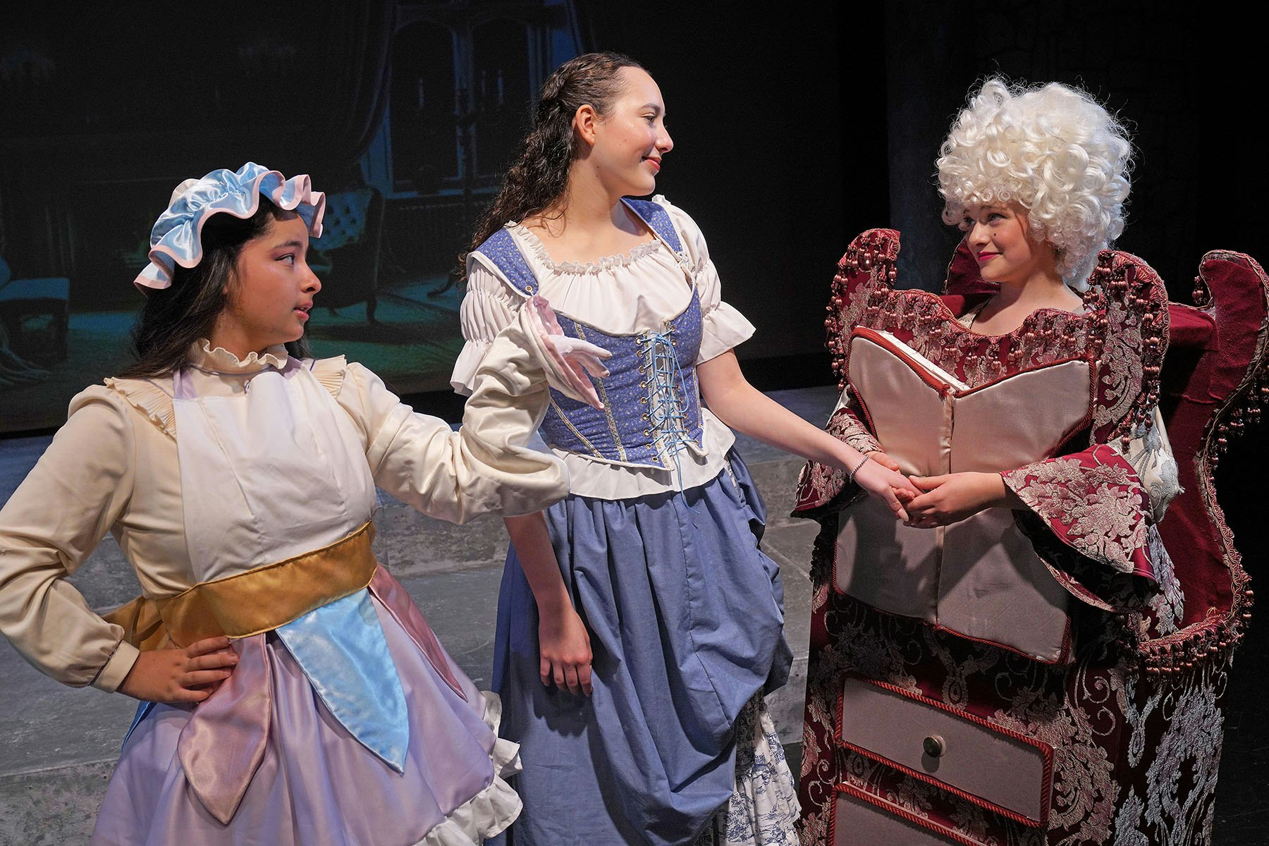 Three people in theatrical costumes, one in a maid outfit, another in a blue dress, and one in a baroque style with a curly white wig.
