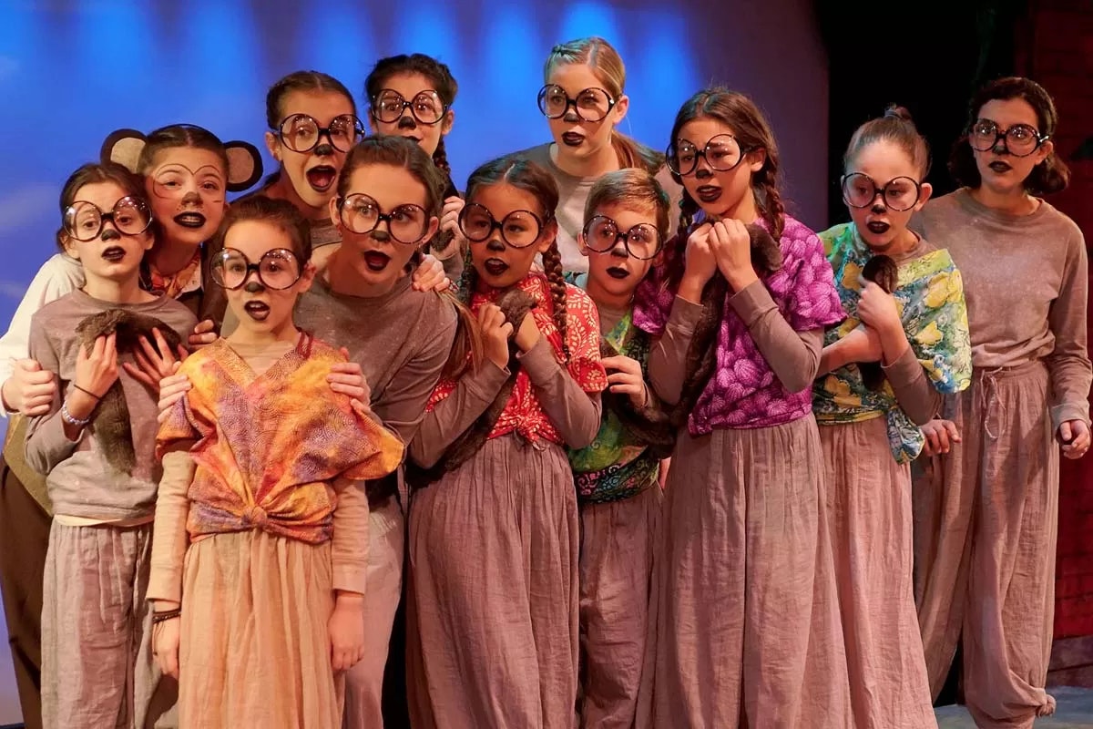 Group of children in costumes with large glasses and dark lipstick, posing on stage with a playful expression.