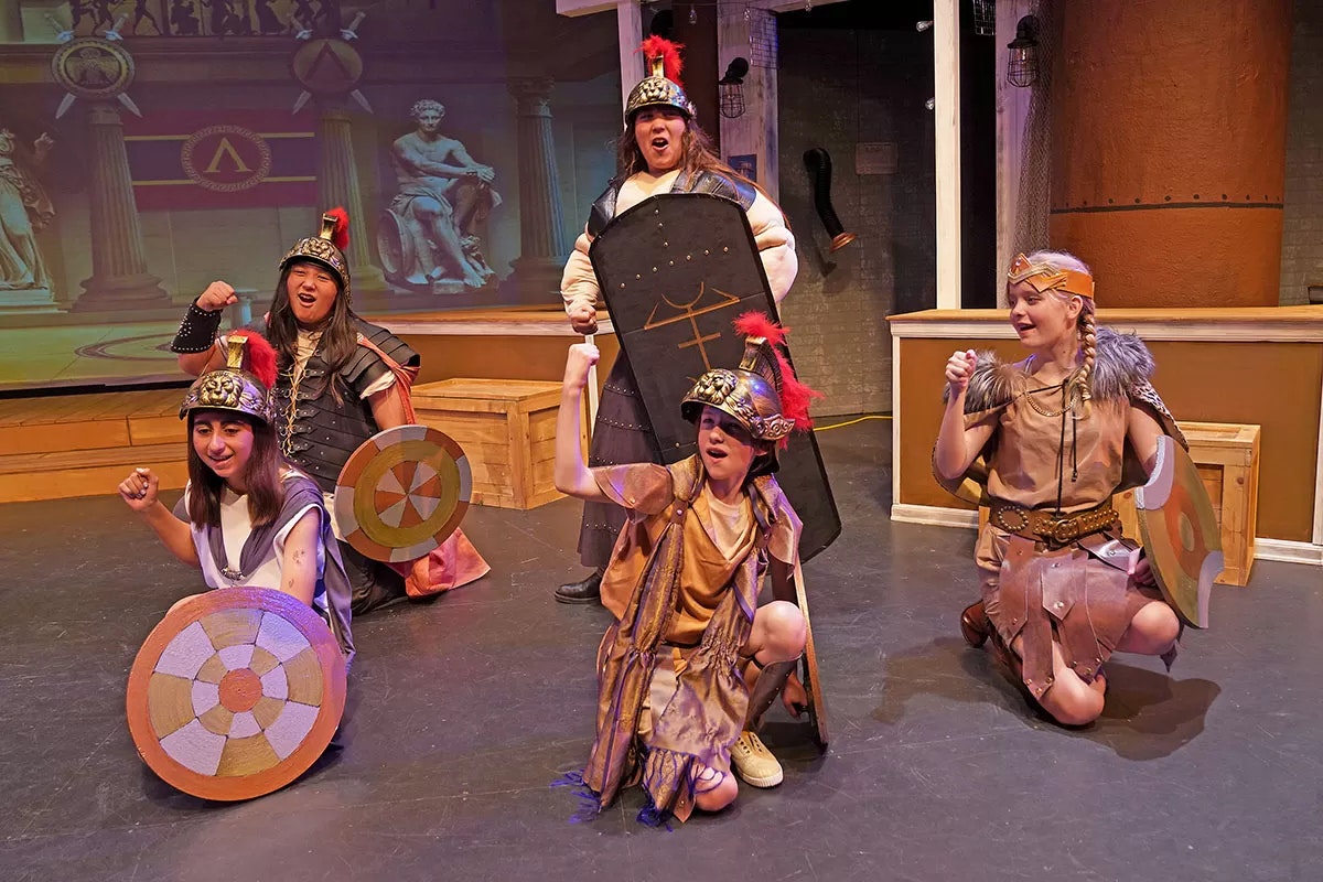 People in costumes with shields on stage, acting in a theatrical play.