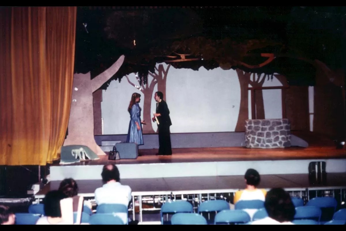 A stage play scene with two actors in costume, an audience watching, and a set resembling a forest with a well.
