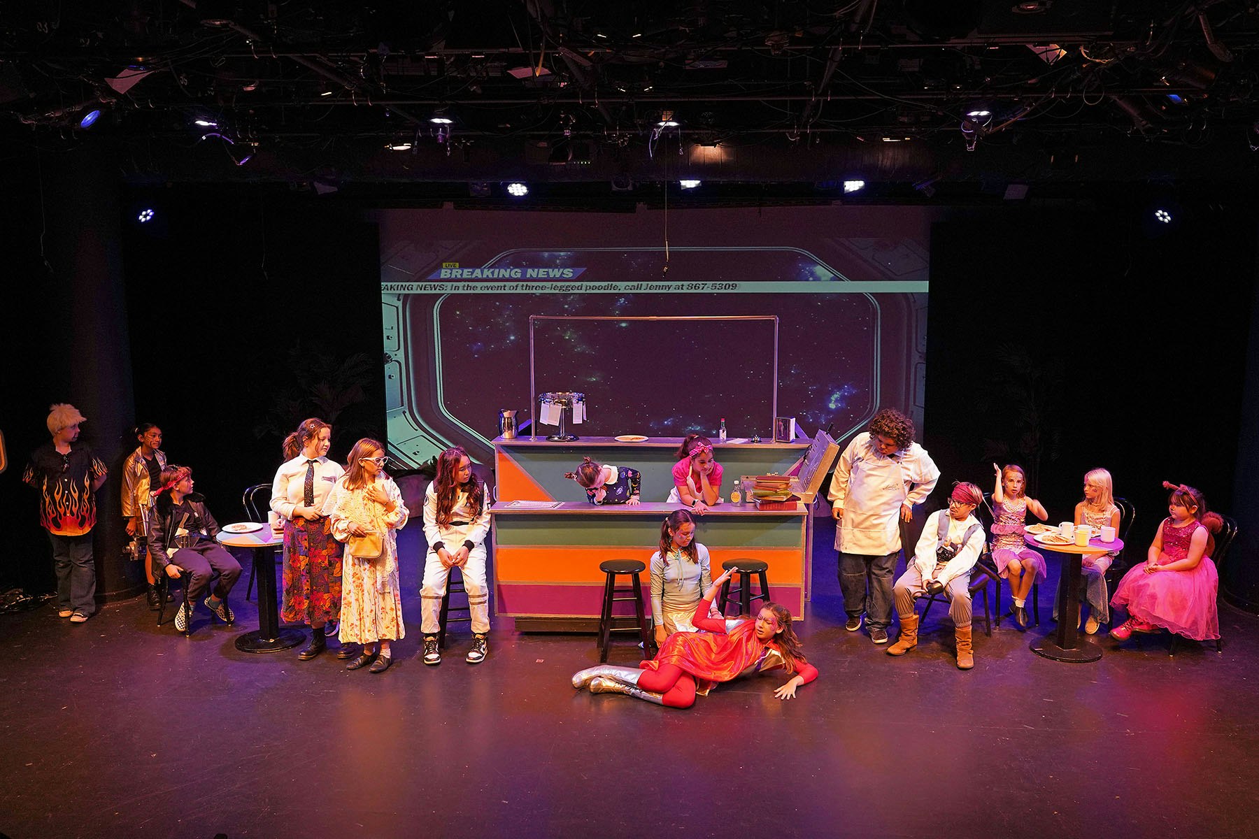 A vibrant theatrical stage with performers in costume, set in a cafe with a "Breaking News" screen backdrop.