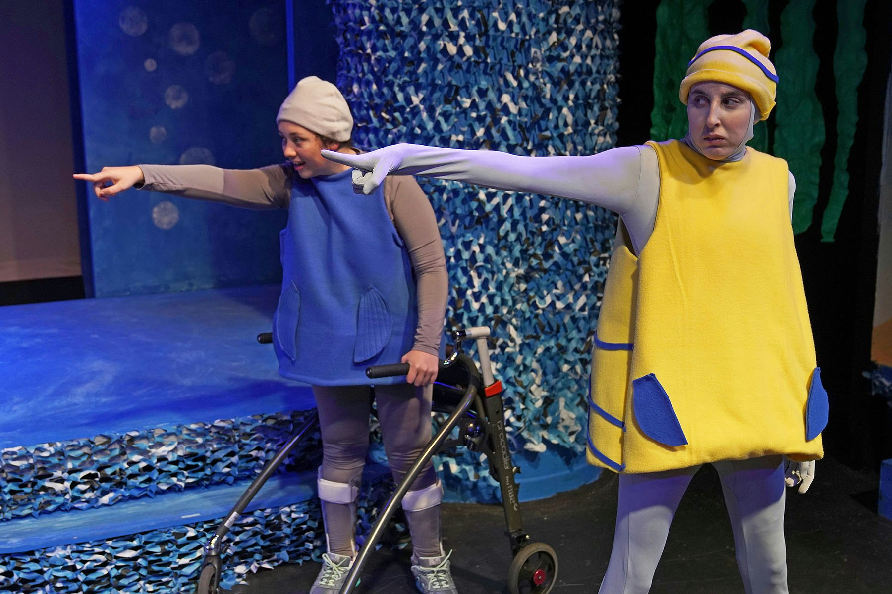 Two people in colorful costumes pointing dramatically on a stage set with a blue, aquatic theme.