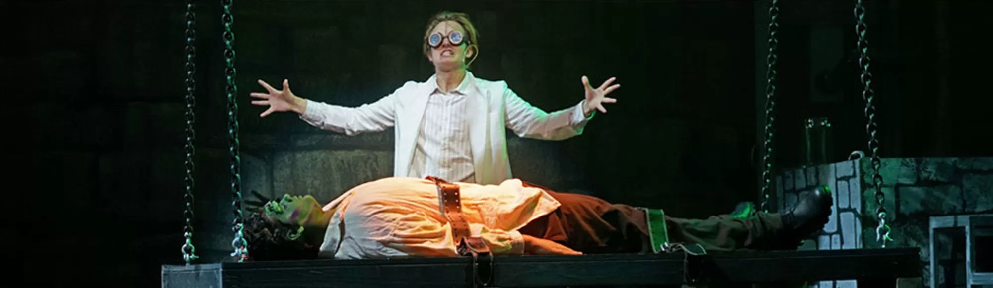 A person in a white coat with goggles raises their arms behind a green-skinned figure strapped to a table, in a dark, theatrical setting.