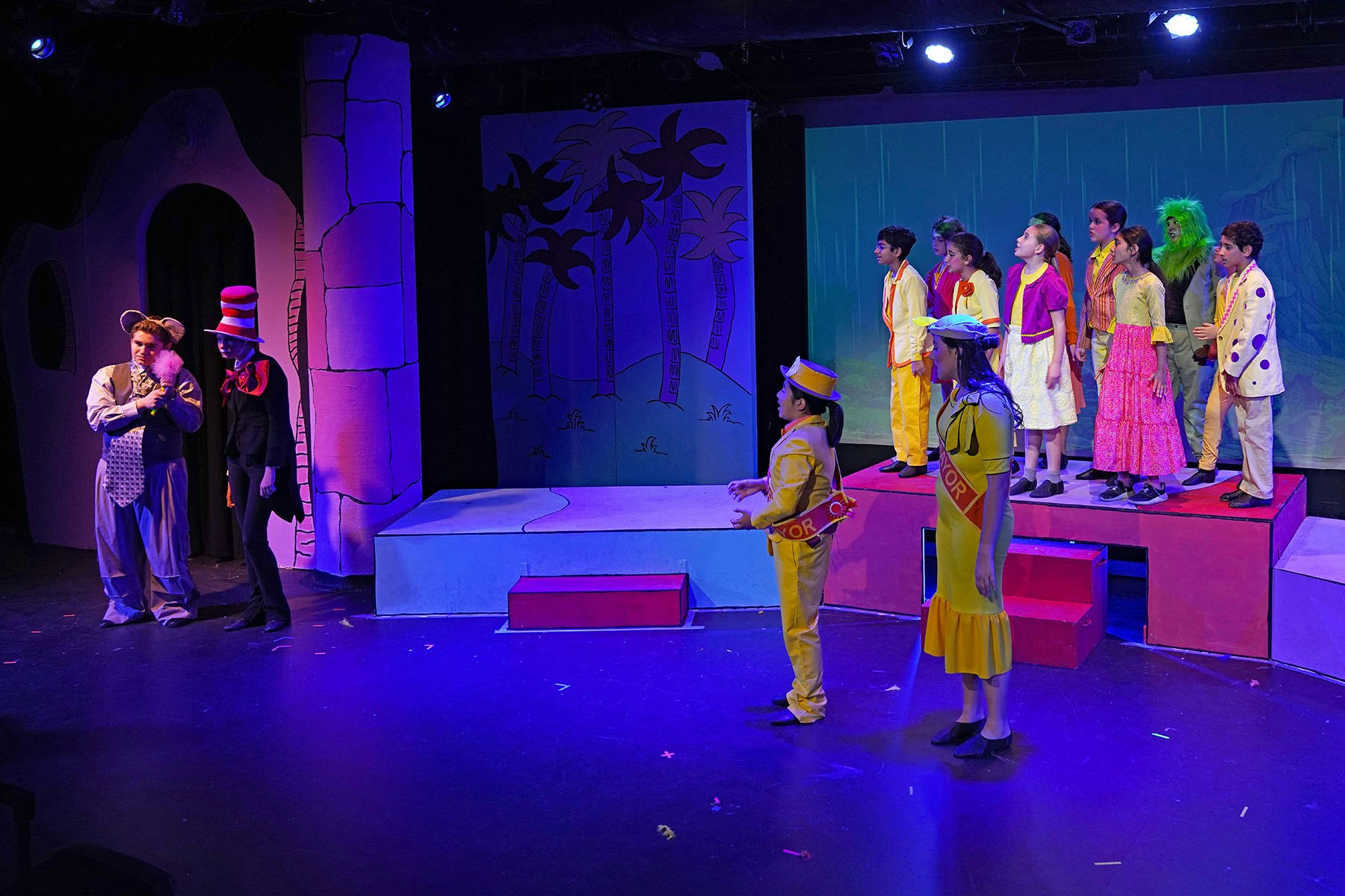 A colorful stage scene with actors in costumes, performing for an audience, featuring characters like a cat and a mouse.