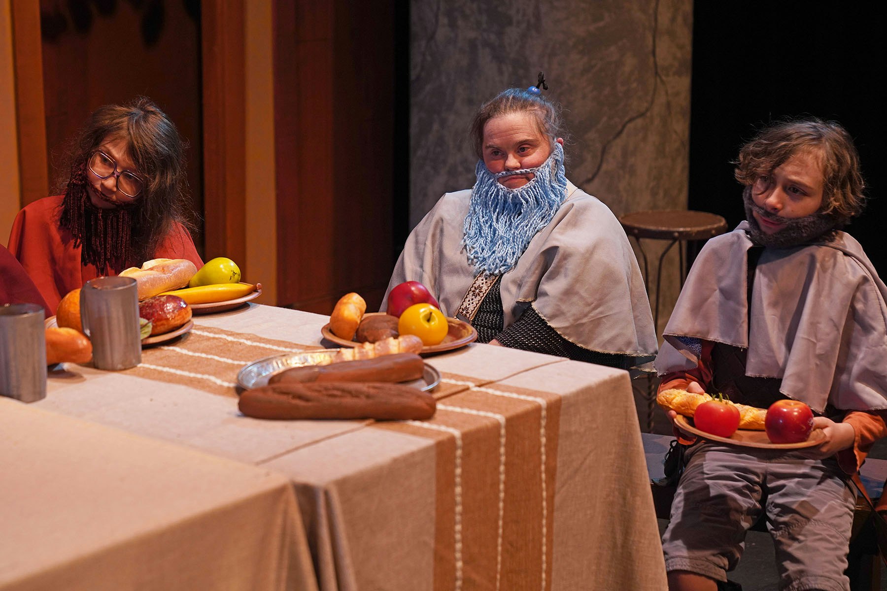 Three people in costume with fake beards sit at a table set with plastic food and drink.