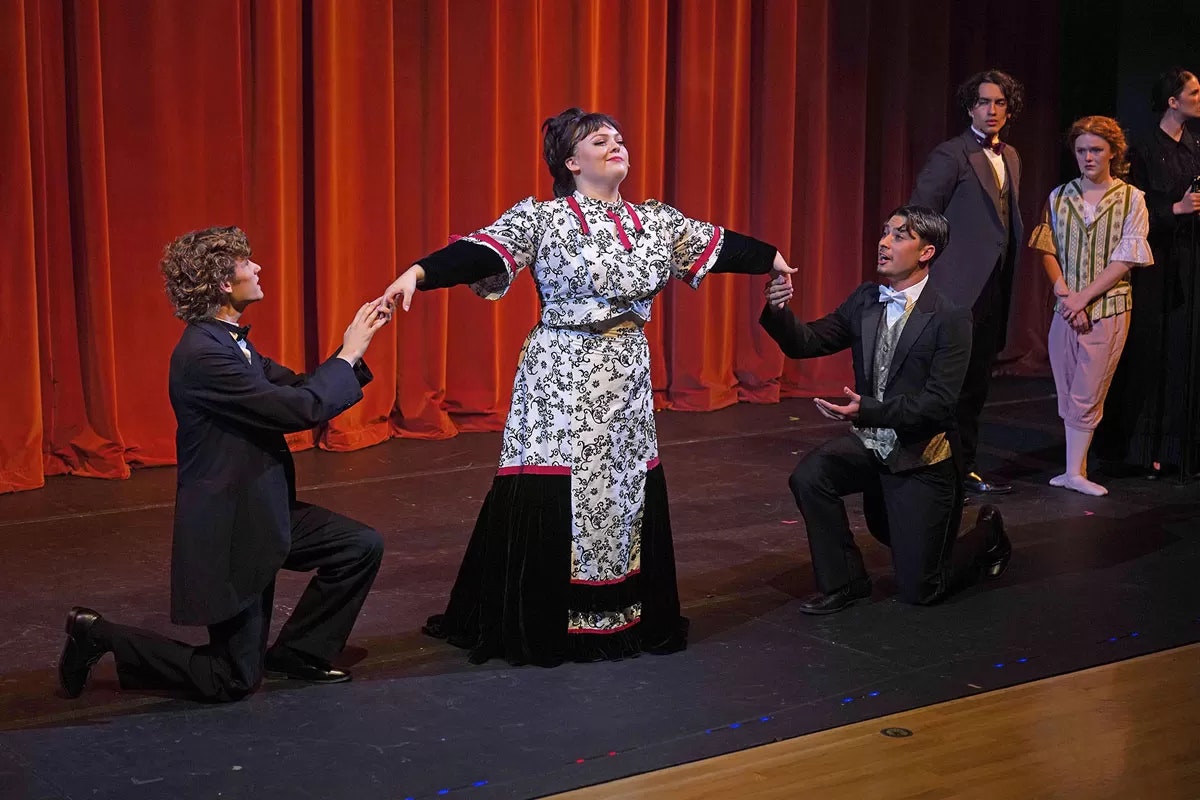 A theatrical scene with actors in period costumes on stage, two kneeling before a central figure, with a red curtain backdrop.