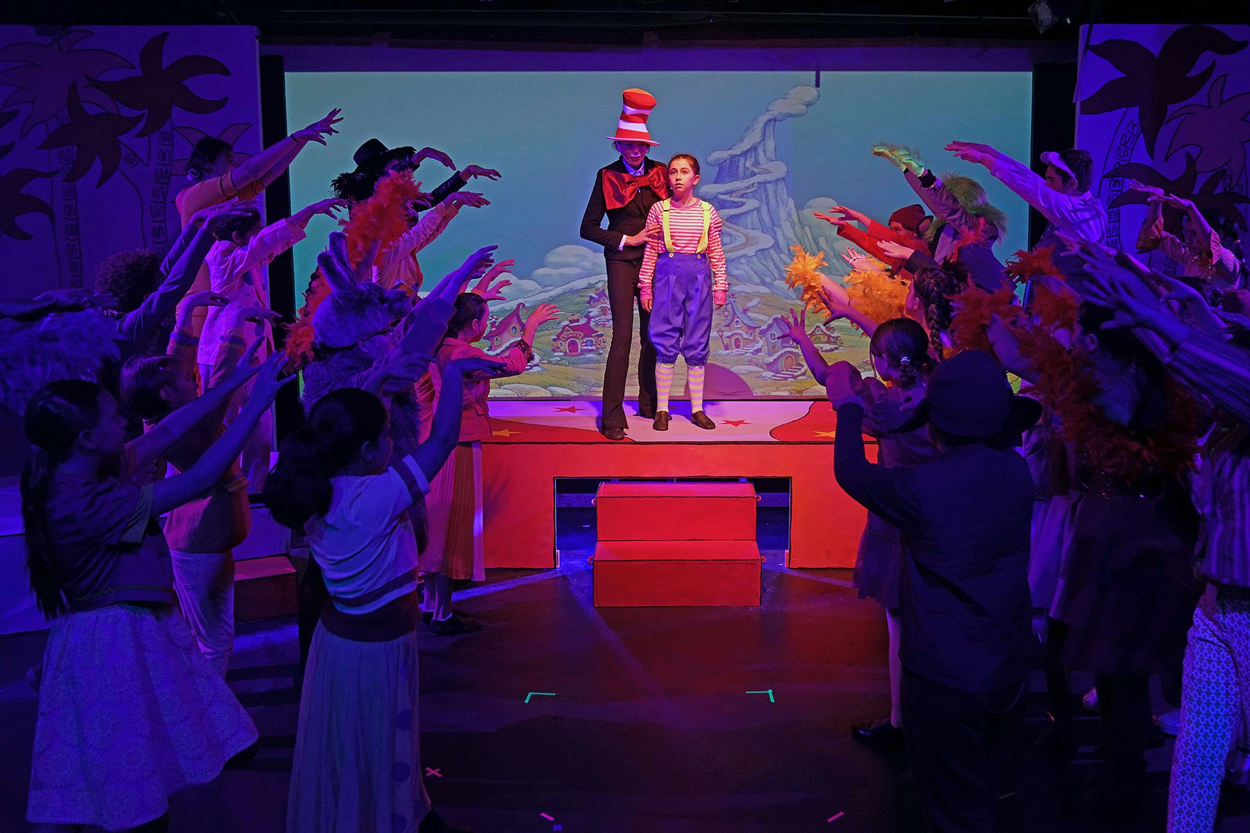 A colorful stage performance with actors in costumes, featuring a central character and a lively ensemble in an animated setting.