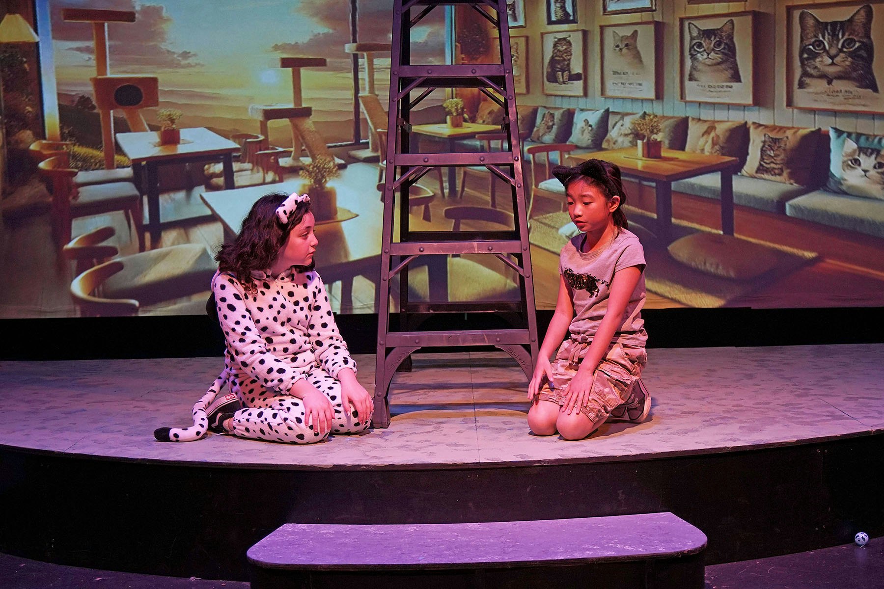 Two people in animal-themed costumes are sitting on a stage with a backdrop of cat decor and furniture.