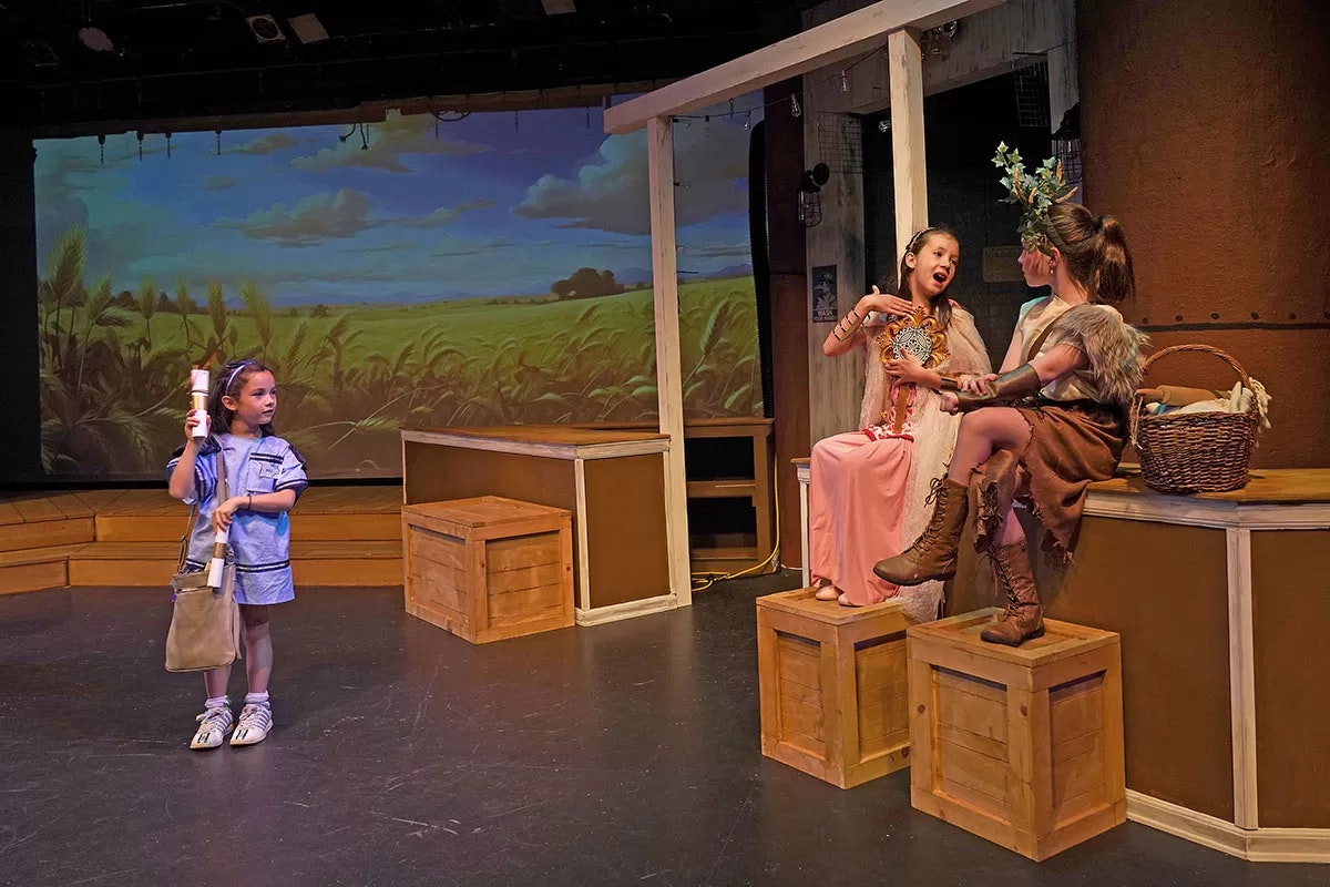 Children performing a play on stage; one holds scrolls while two sit on crates, one with a flower crown and a basket nearby.