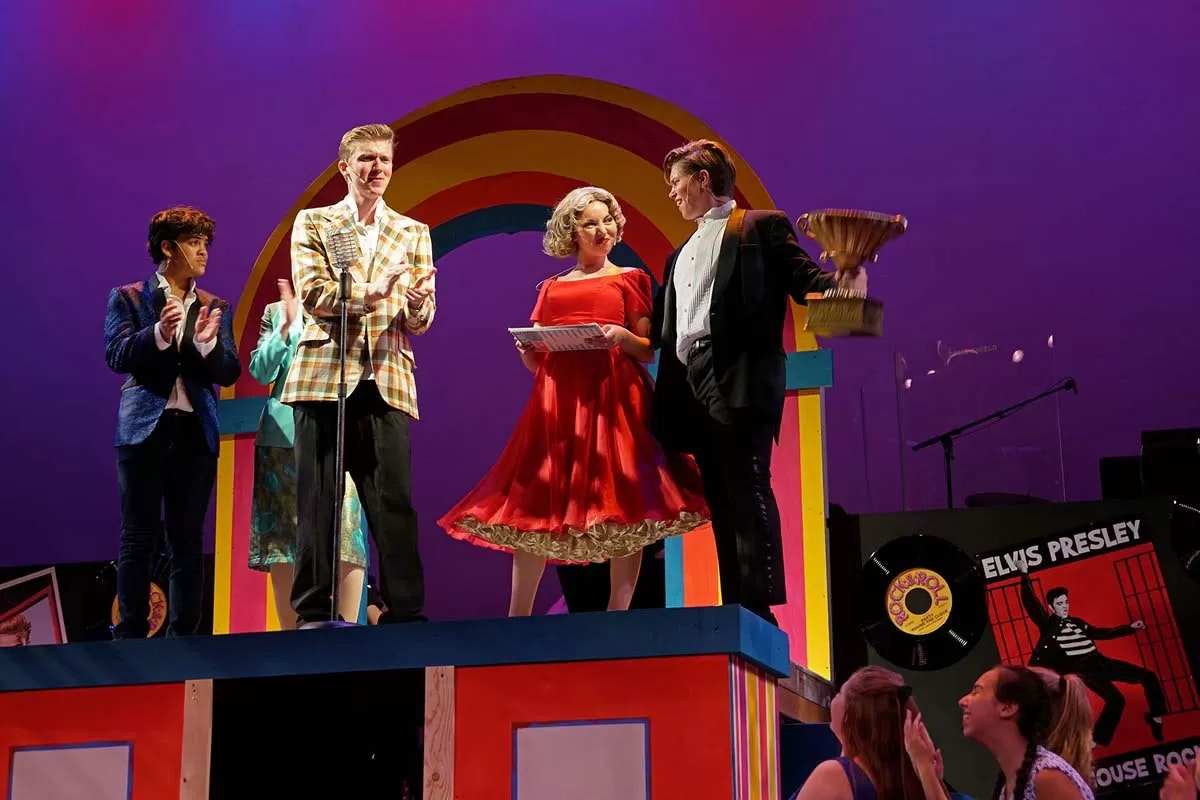People on stage during a colorful performance, one holding a trophy. Elvis Presley poster in the background.