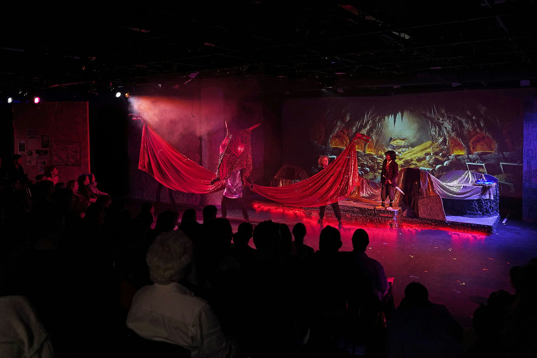A theatrical stage with actors, red fabric props, and a backdrop of a cave, viewed by an audience.