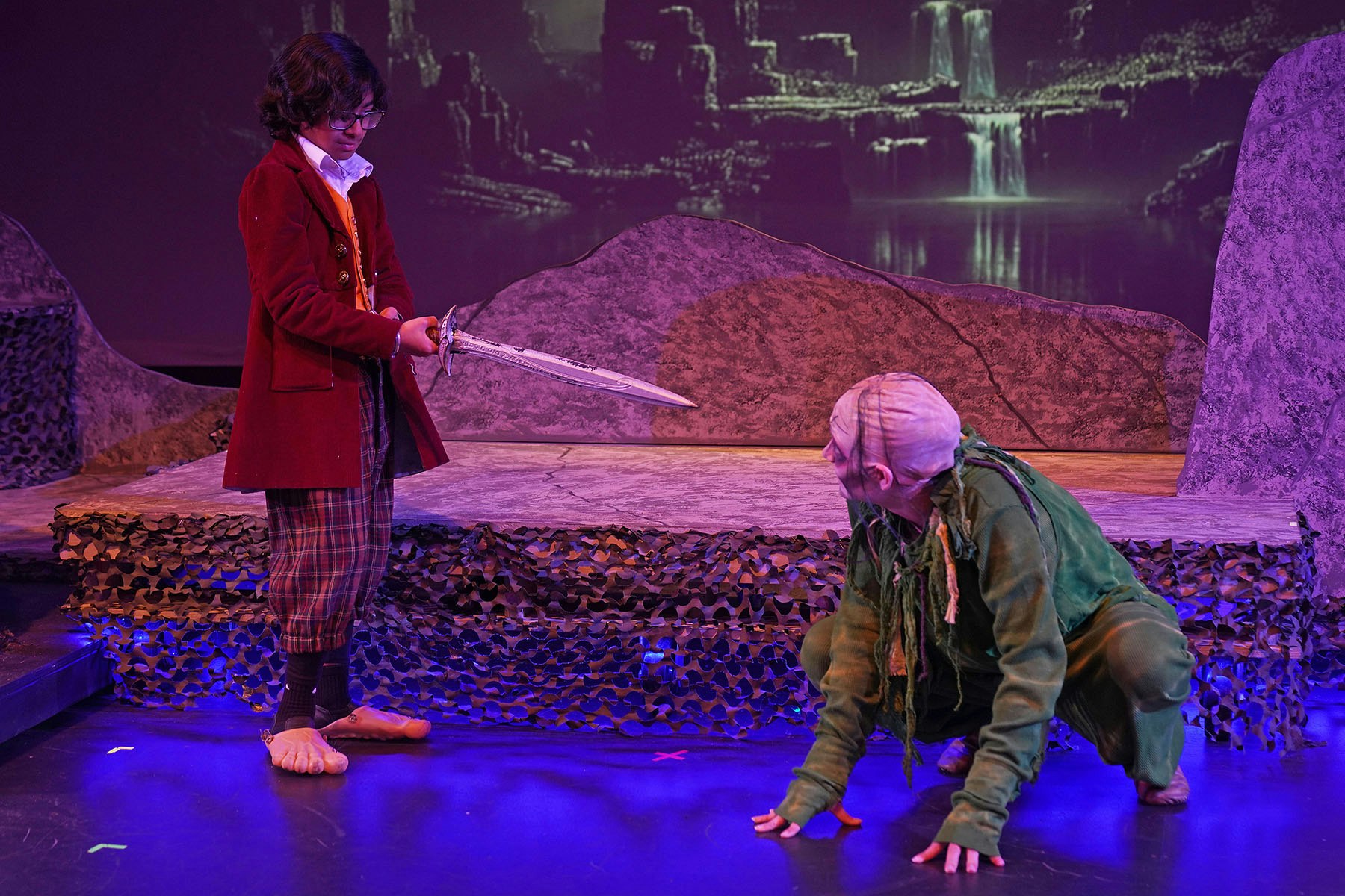 Two actors on stage, one with a sword in a fantasy setting with rock-like props and a waterfall backdrop.