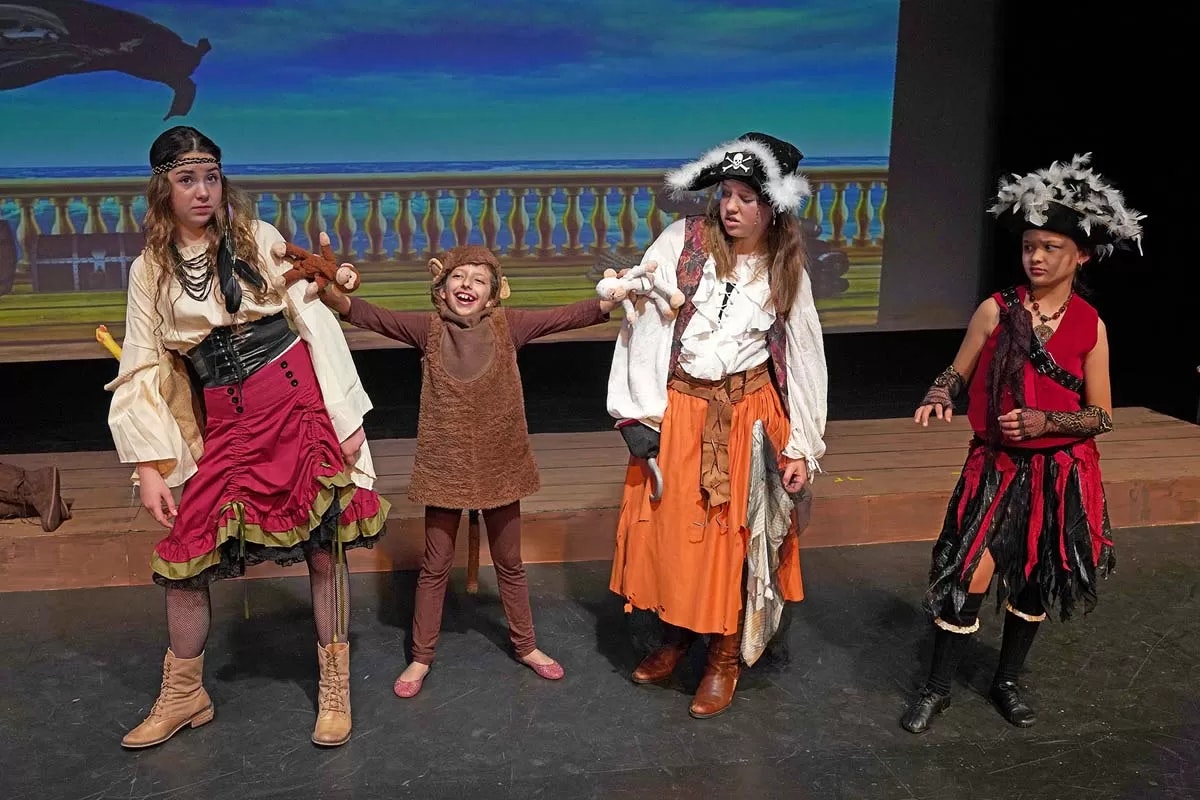 Four people in pirate and monkey costumes on stage, acting out a scene with a ship backdrop.