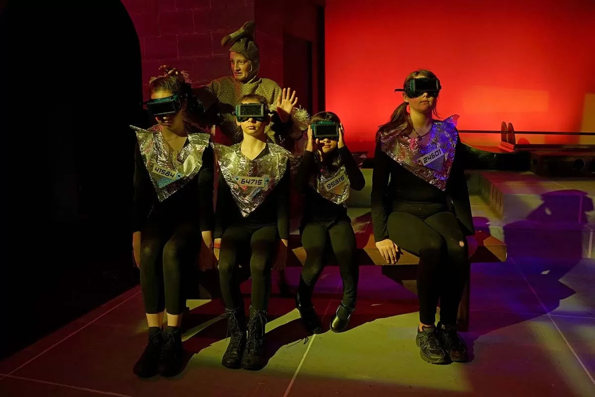 Four people in futuristic outfits and goggles sit on a bench; another person behind them gestures with hands.