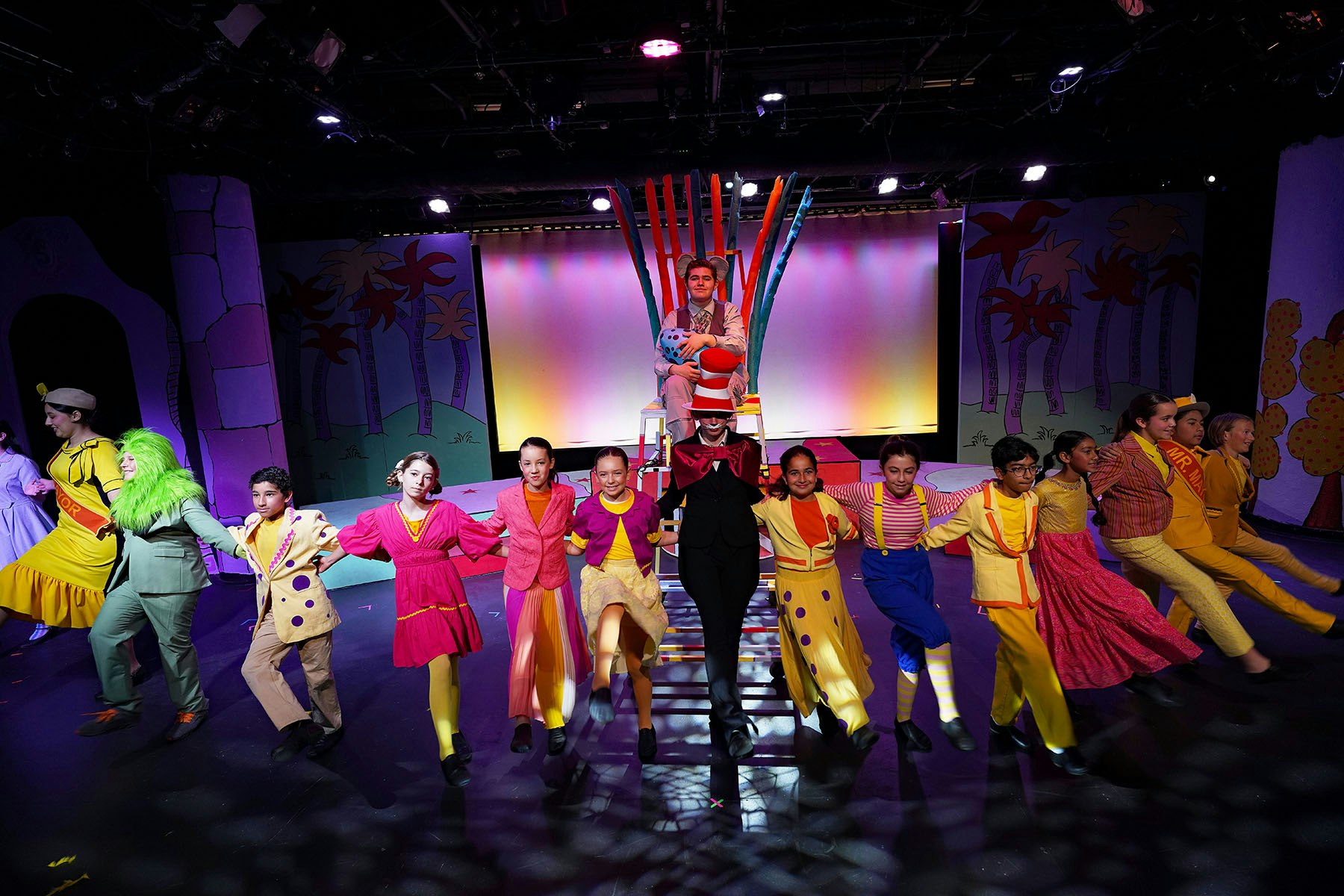 A vibrant stage performance featuring actors in colorful costumes, dancing and posing together in a lively scene.