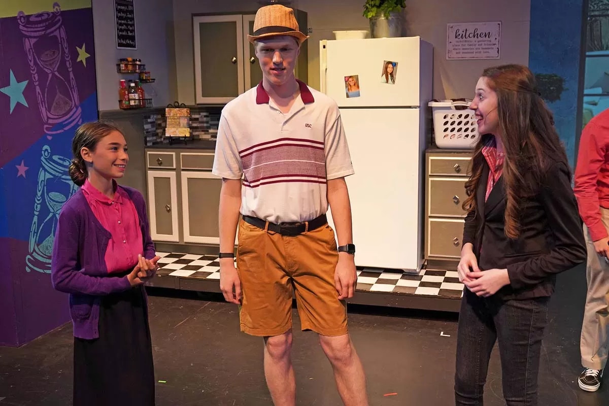 Three people smiling in a kitchen-themed stage setting.