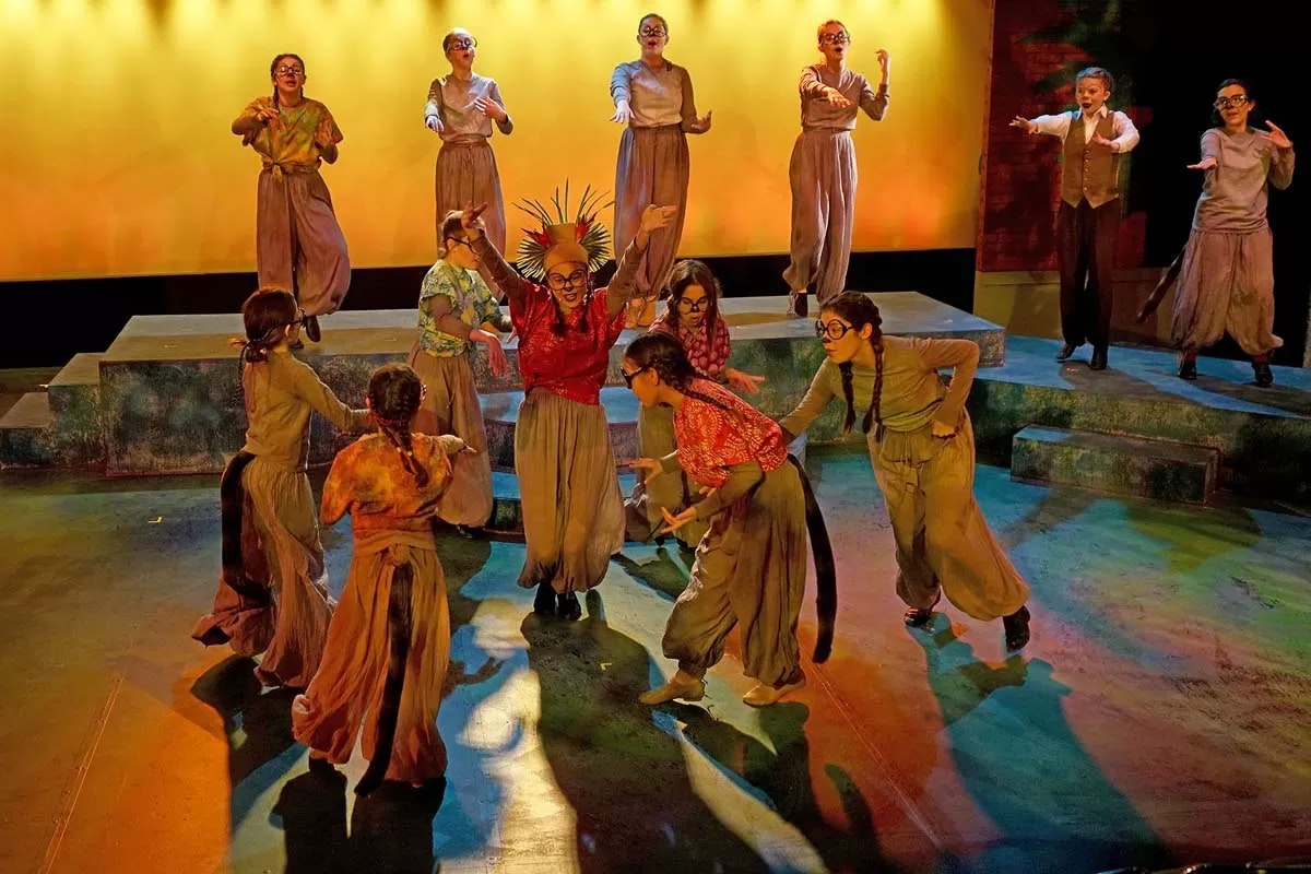 A group of people performing on stage, wearing costumes and dramatic makeup, under colorful lighting.