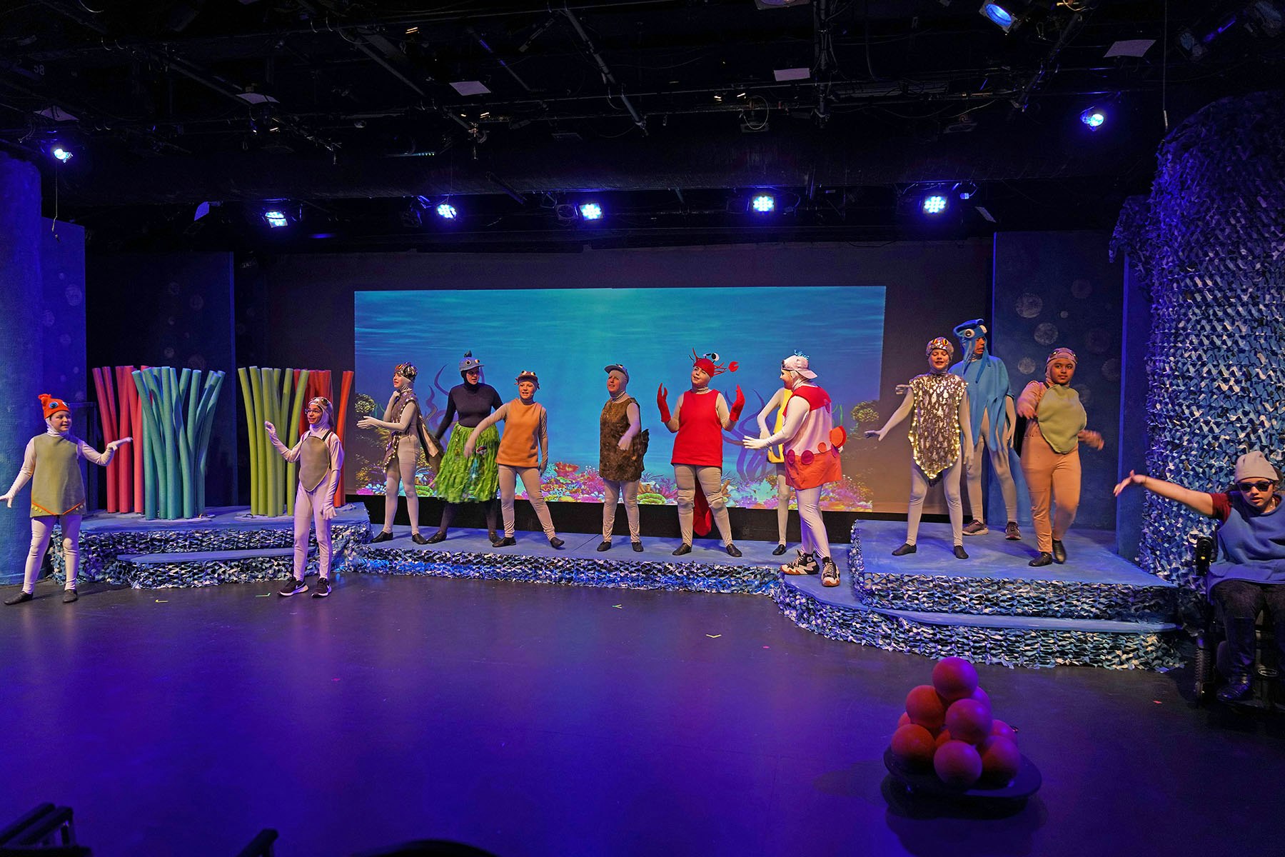 A colorful stage play with performers in ocean-themed costumes against a sea backdrop.