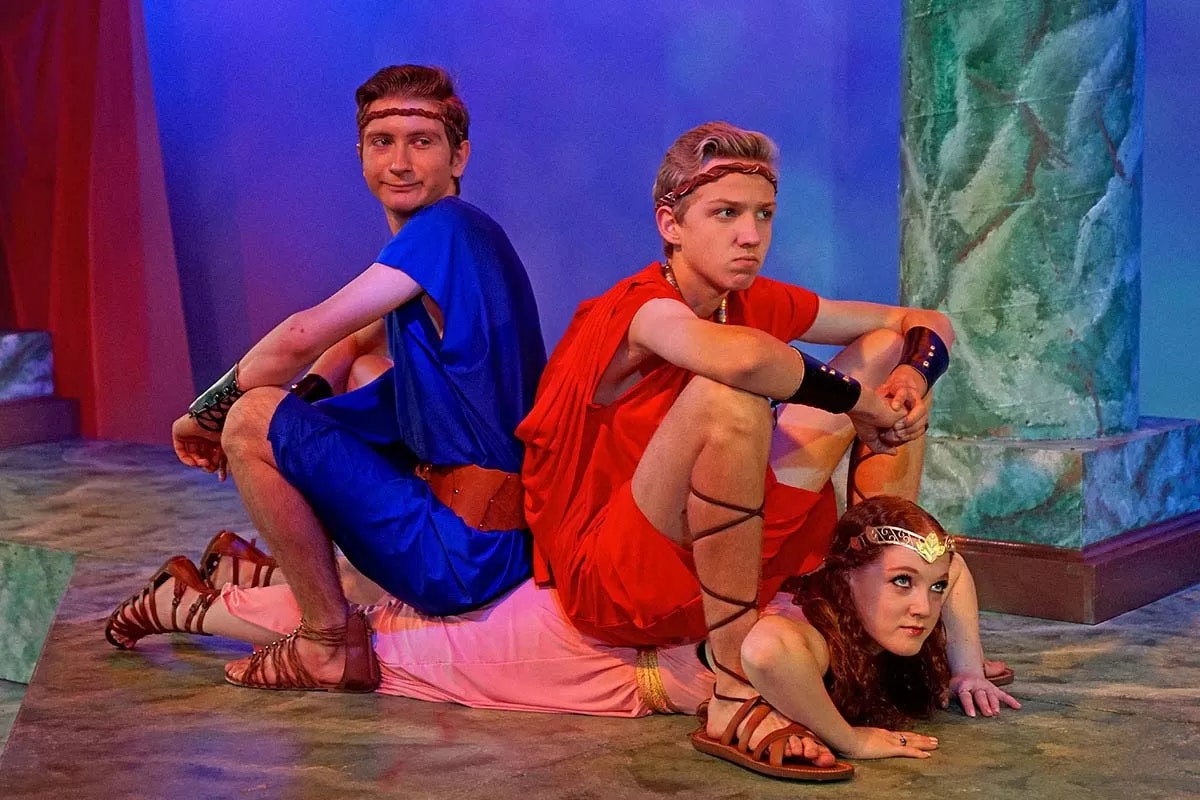 Three people in Greek-style costumes on stage, two sitting with a person lying beneath them.