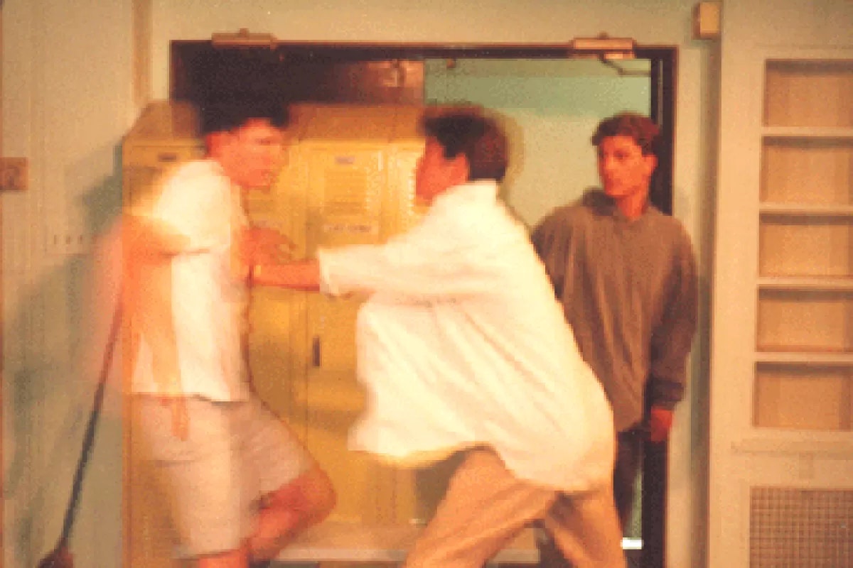 Two people are engaged in a physical confrontation near lockers, with a third person watching.