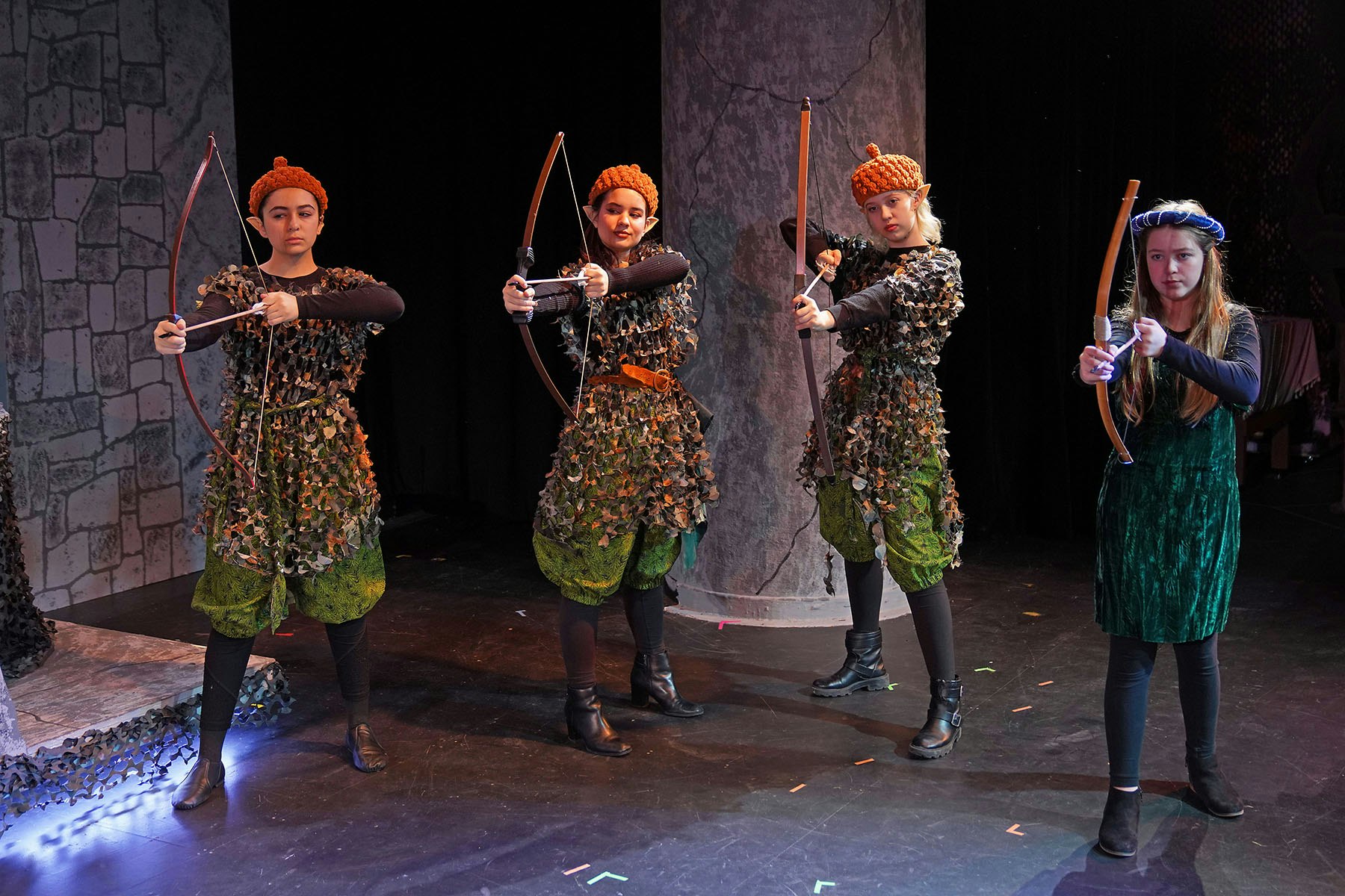 Four people in elf costumes, aiming bows and arrows on stage.