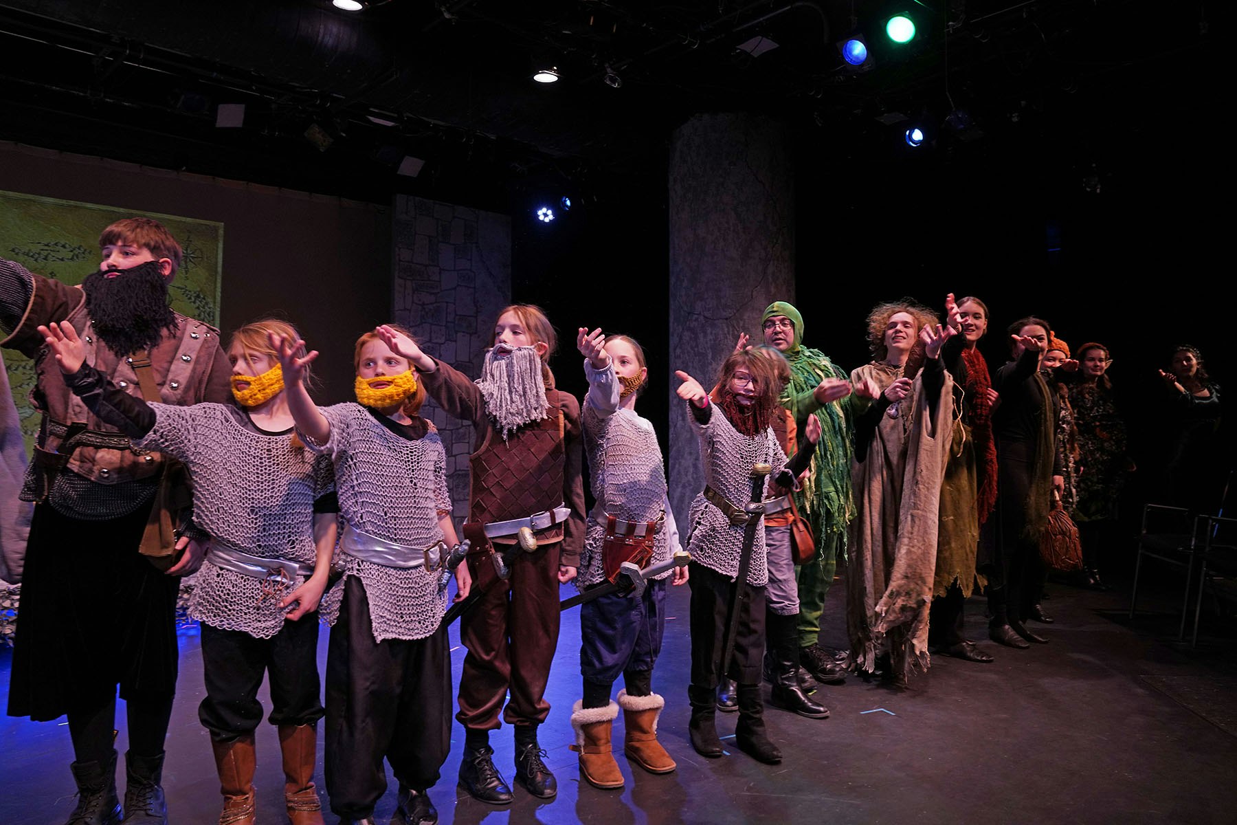 A group of people in costumes with beards and armor perform on stage, reaching forward energetically.
