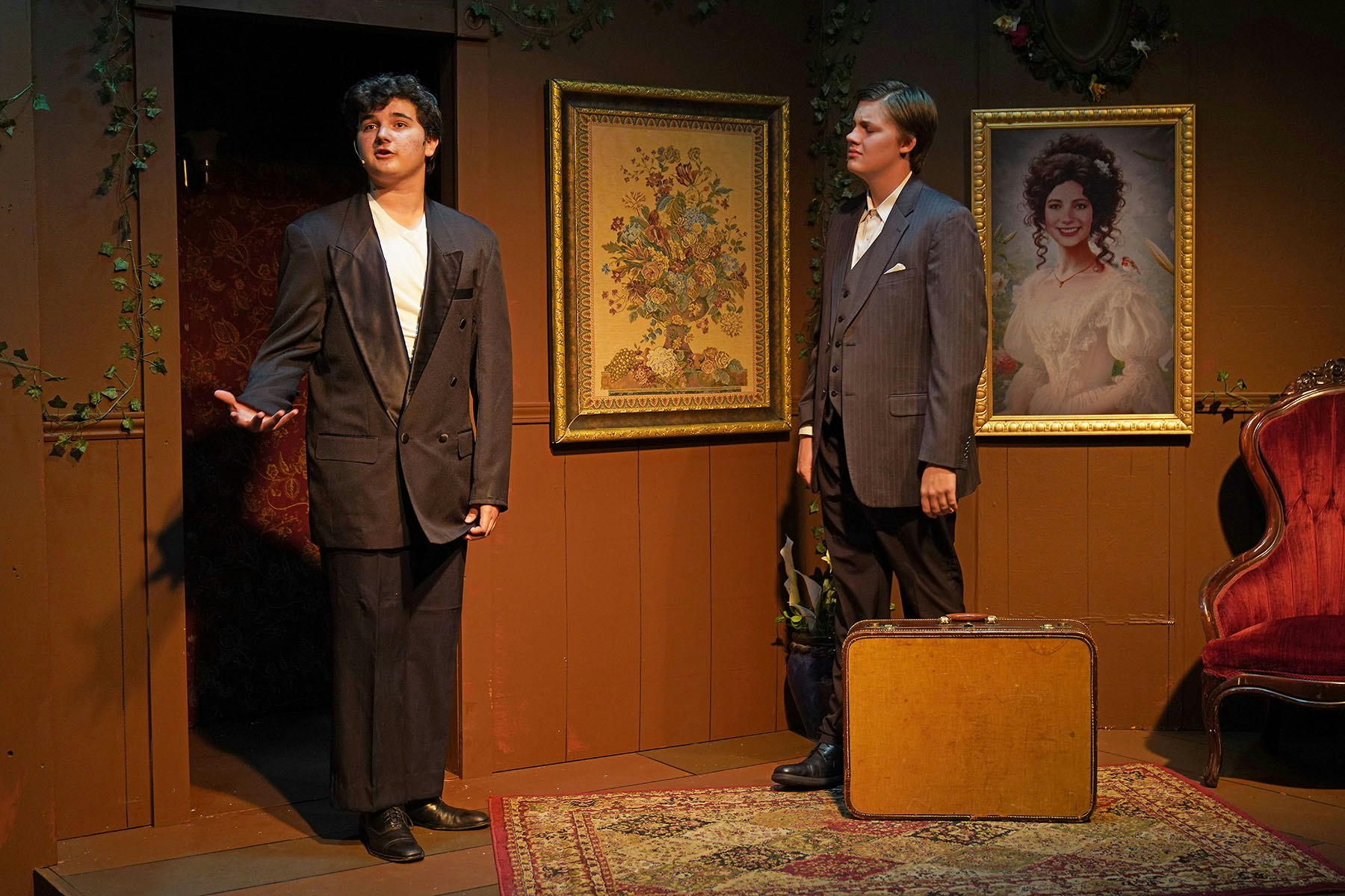 Two men in suits in a room with paintings, a suitcase, and a red velvet chair.