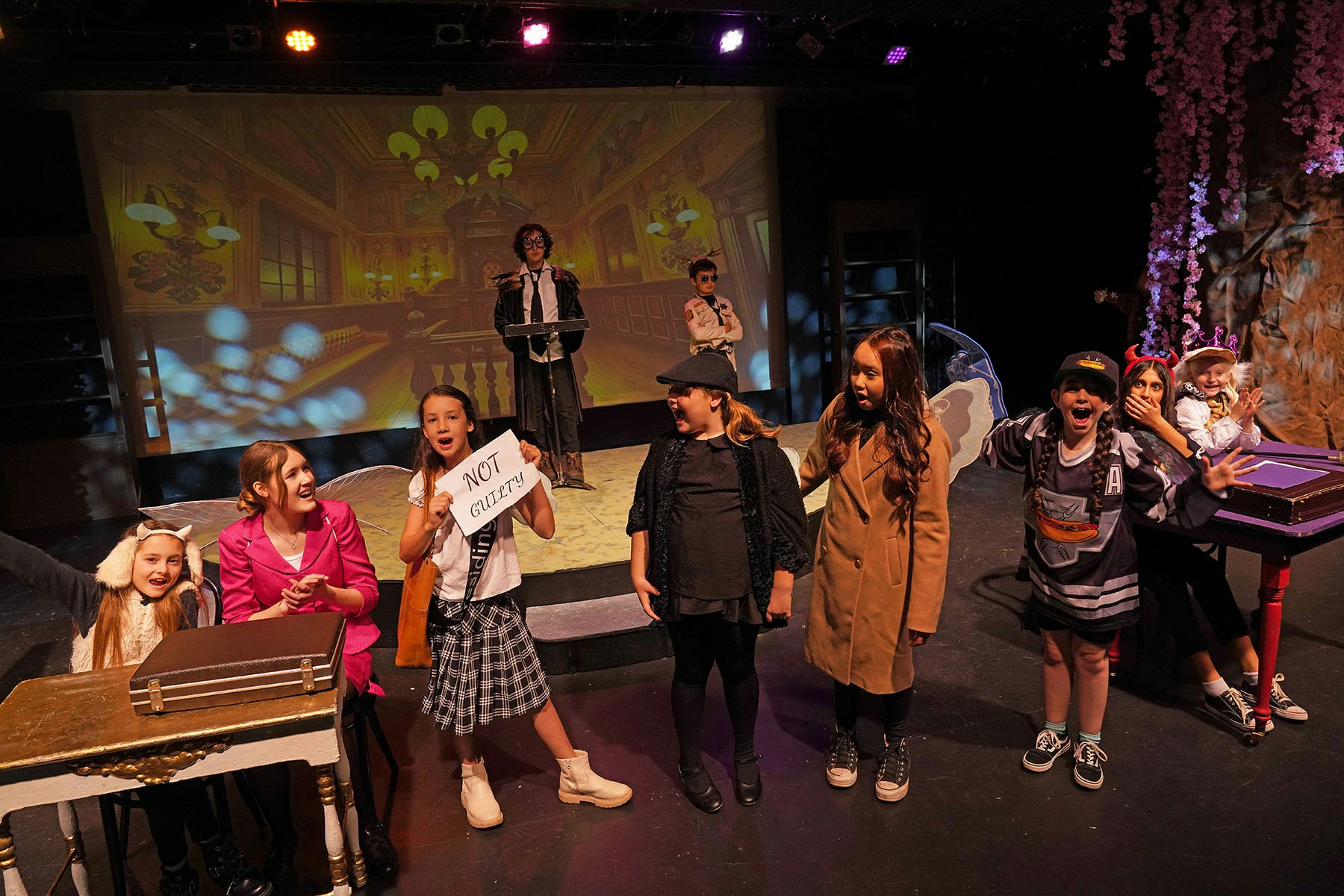 A group of children acting in a play, one holds a "NOT GUILTY" sign, with a courtroom scene projected behind them.