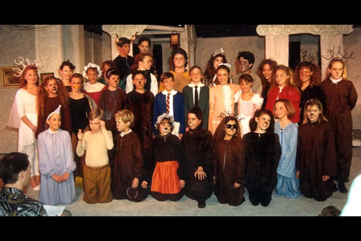 A group of children in costume pose for a school play, with a variety of colorful outfits and accessories like antlers and hats.
