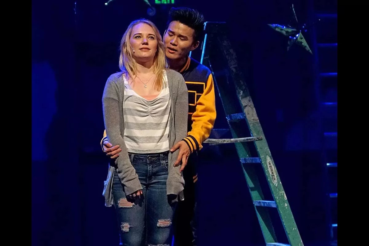 A man wearing a varsity jacket embraces a woman in a striped shirt and cardigan on stage with a ladder in the background.