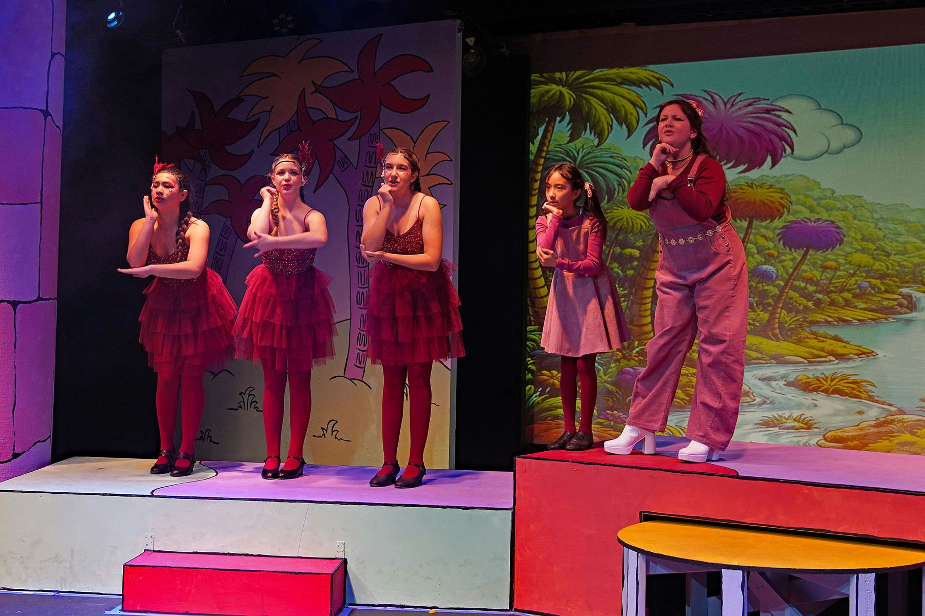 A stage performance featuring five girls in red costumes and one in pink, with a colorful backdrop of palm trees.